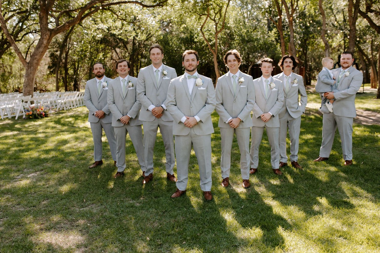 Wedding party in light gray suits standing in formation on a sunny lawn with trees in the background.