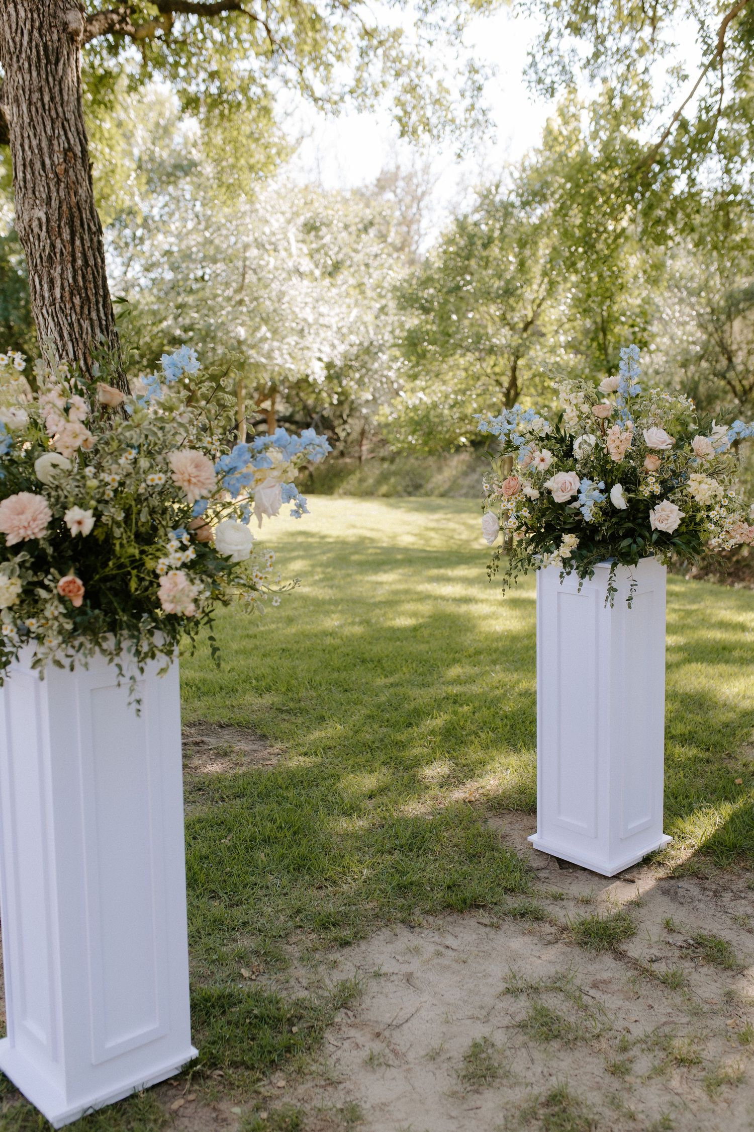 White decorative pedestals with floral arrangements placed outdoors under trees for a wedding ceremony.