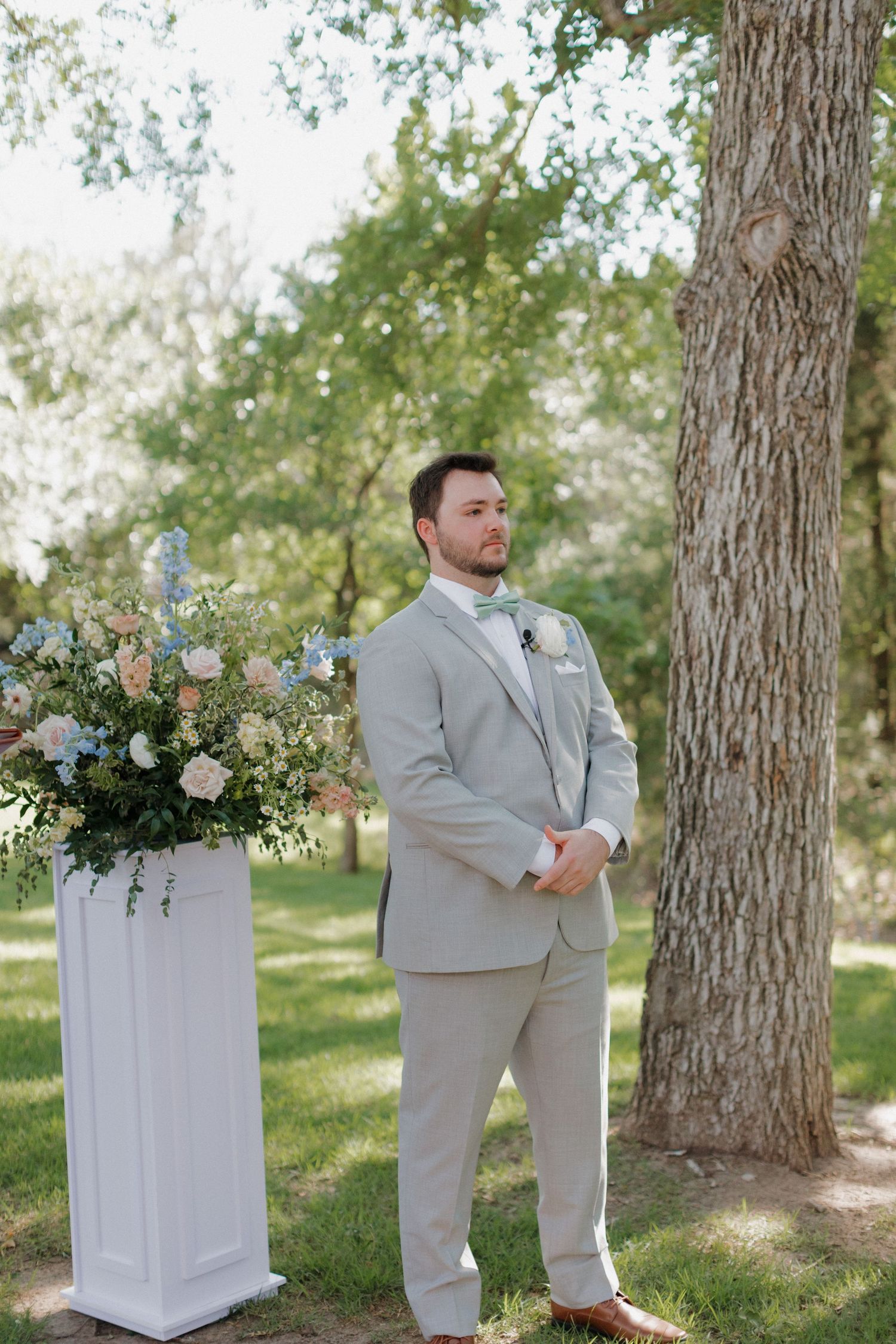 A person in a light gray suit stands beside an elegant floral arrangement and tree trunk outdoors.