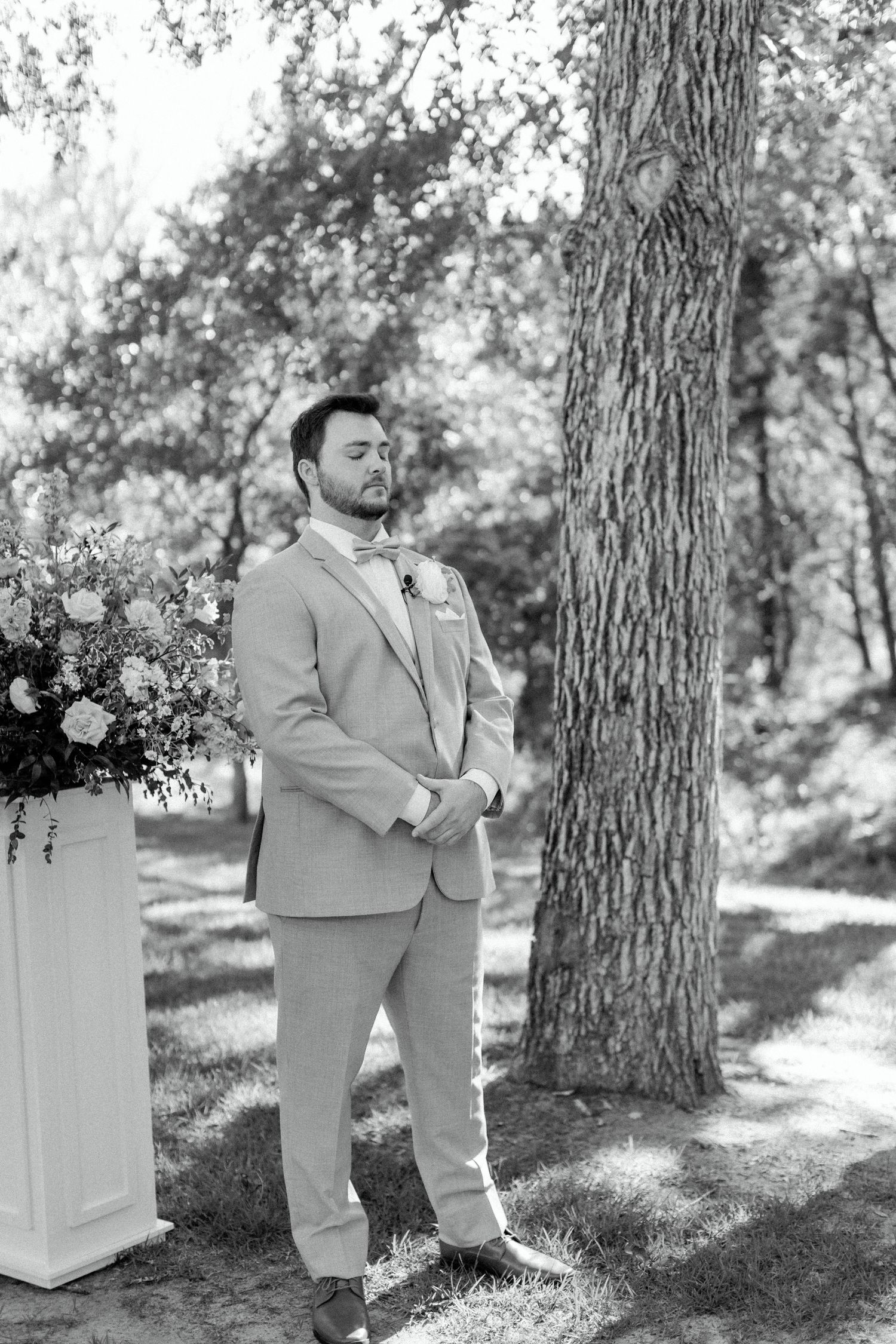 Black and white portrait of a person in a light suit standing next to a floral arrangement outdoors.