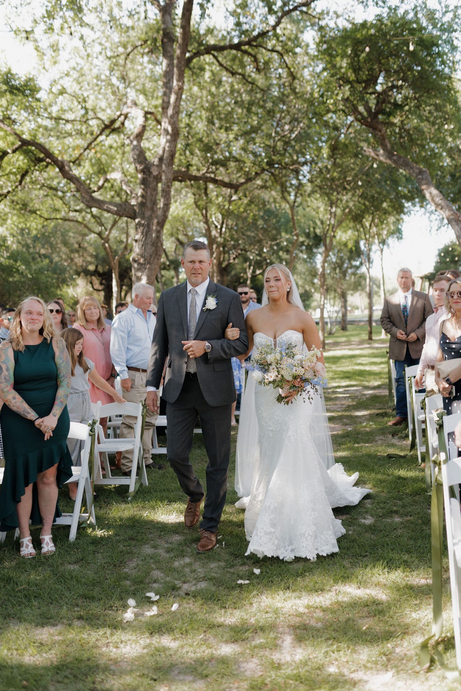 Wedding ceremony processional takes place on grassy path beneath towering trees.