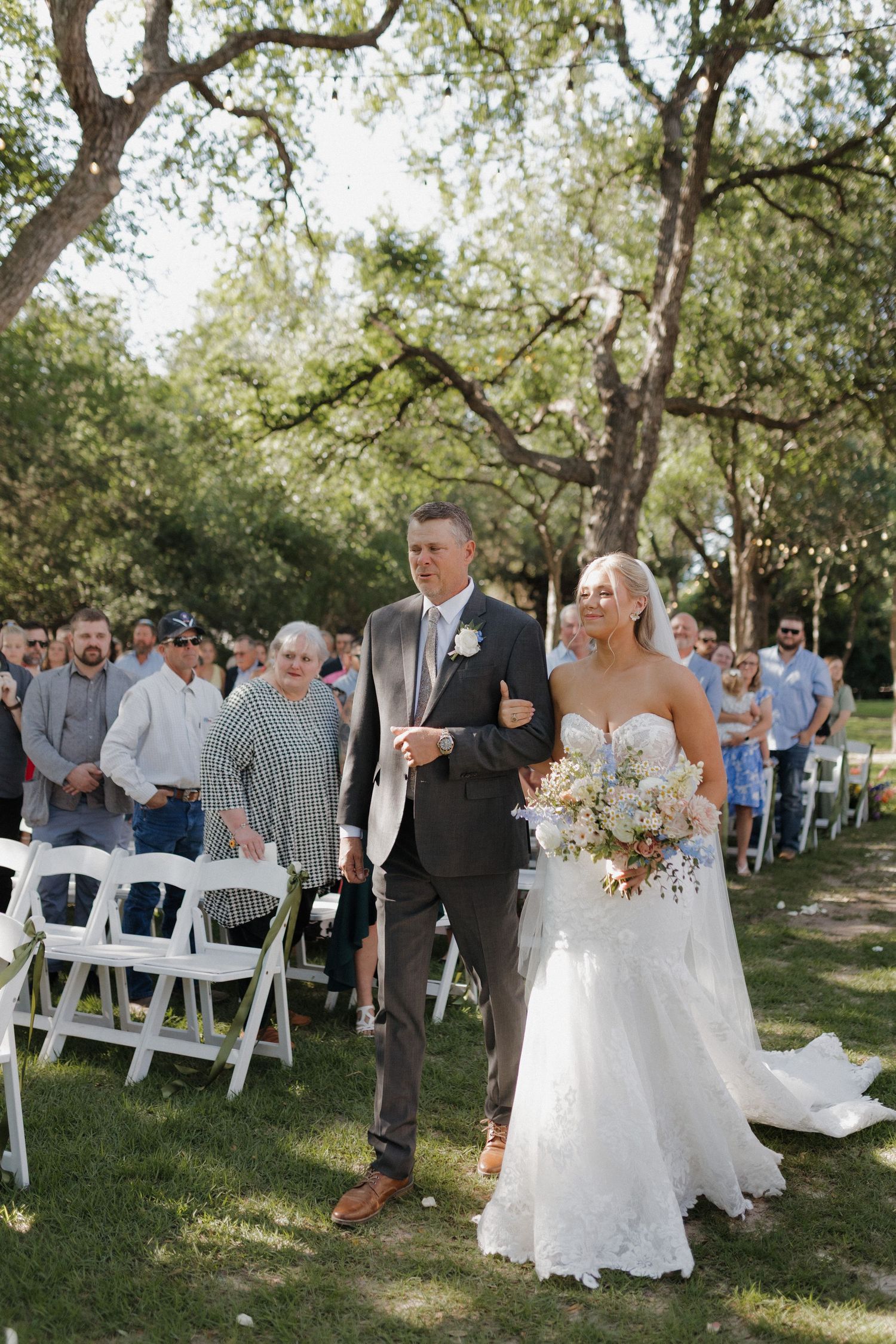Outdoor wedding ceremony setup with white chairs and guests under shady oak trees.