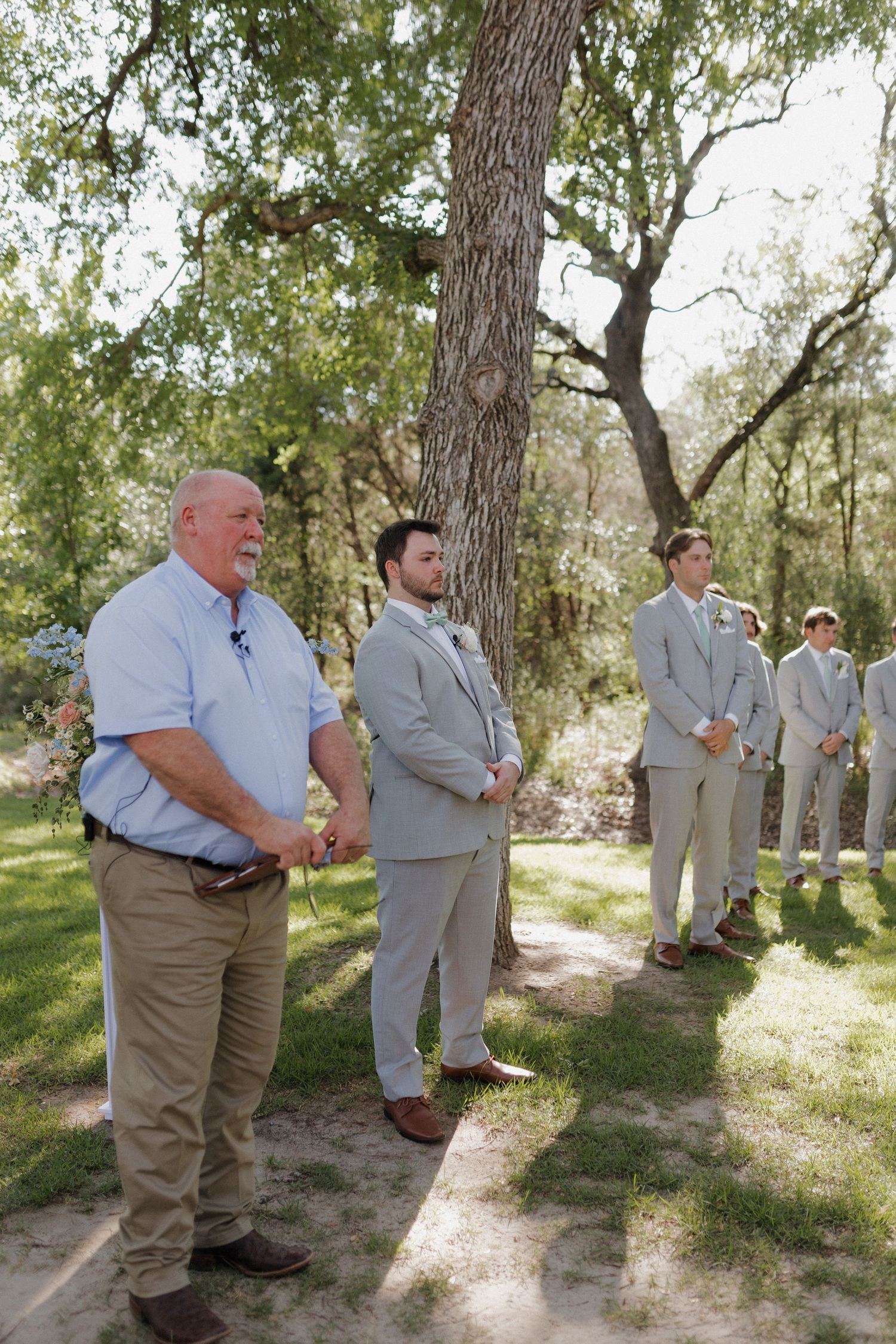 Groomsmen in light gray suits and khaki pants standing outdoors under large oak trees.