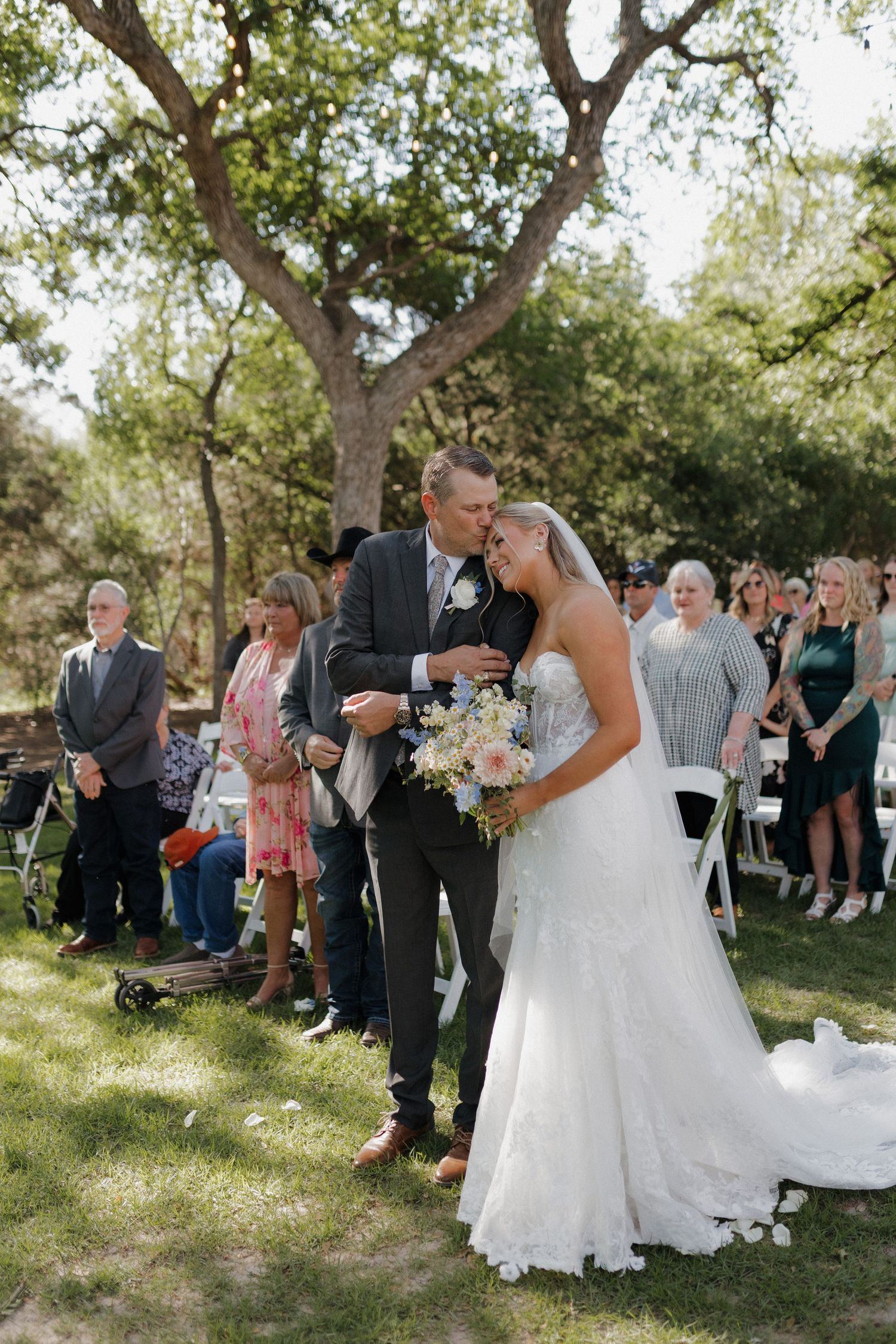 An outdoor wedding ceremony under trees with guests gathered to watch the couple share a special moment together.
