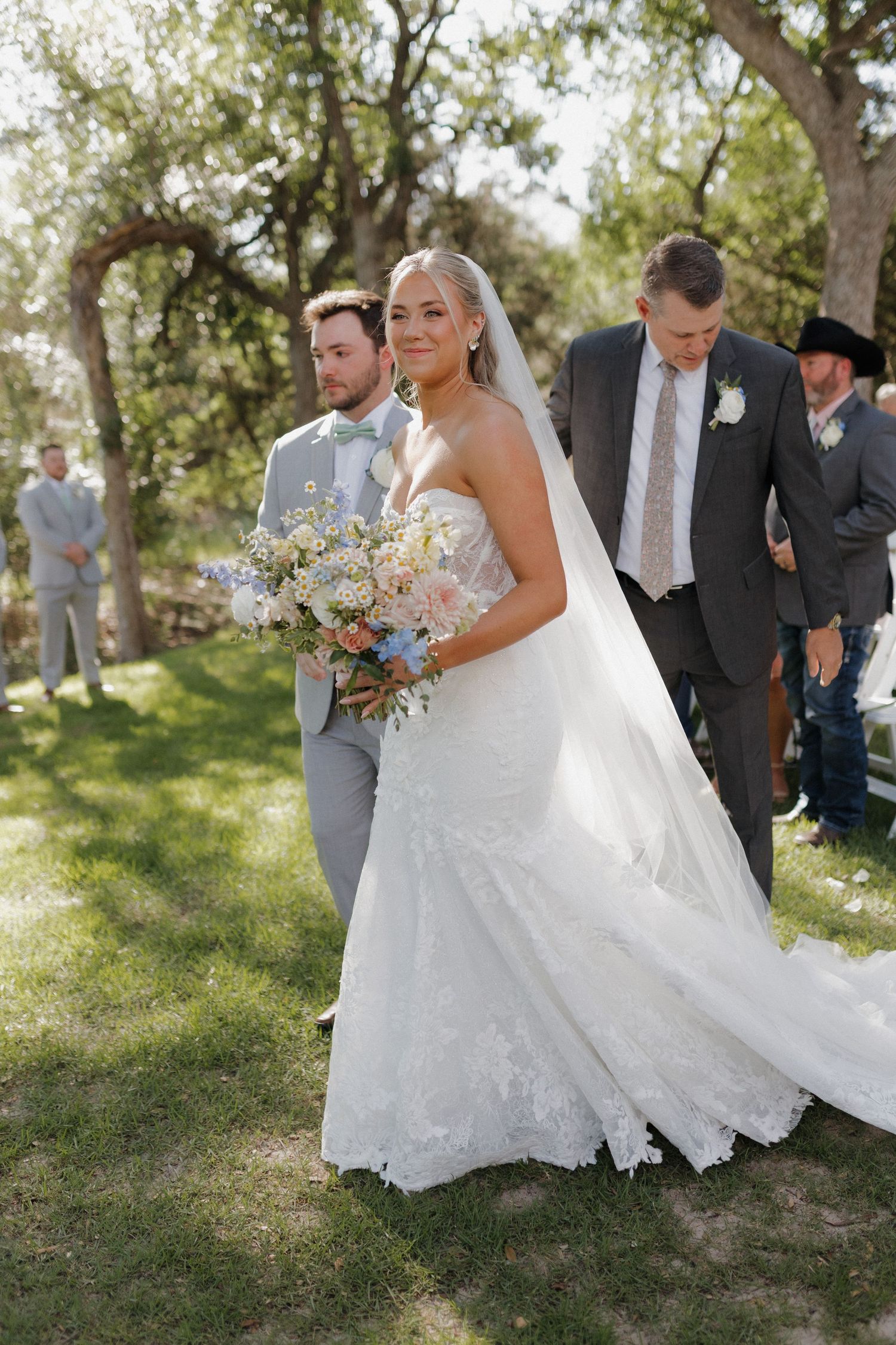 Outdoor wedding ceremony with wedding party walking through a lush tree-lined garden setting.