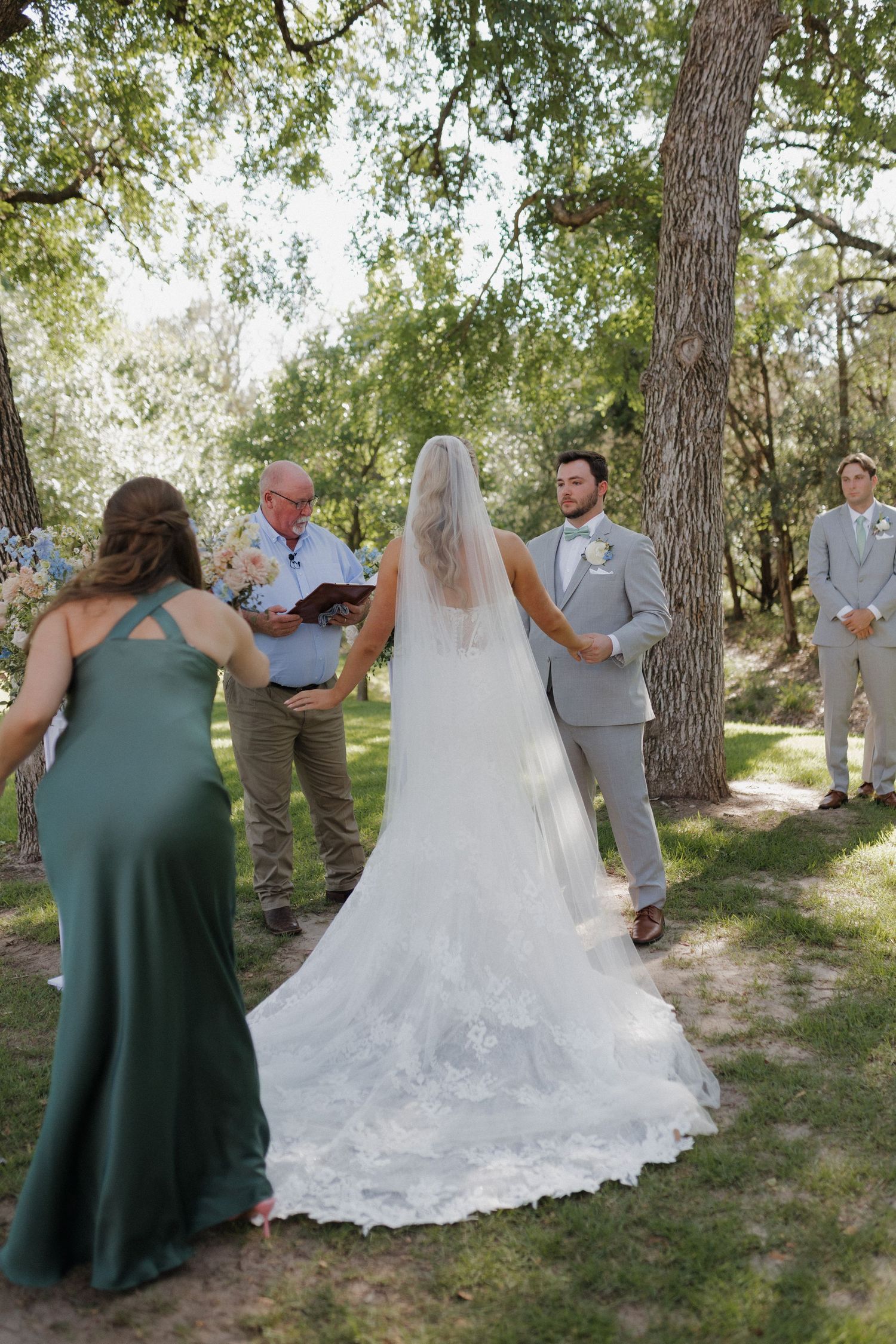 An outdoor wedding ceremony under tall trees with a bride in a lace gown and flowing train.