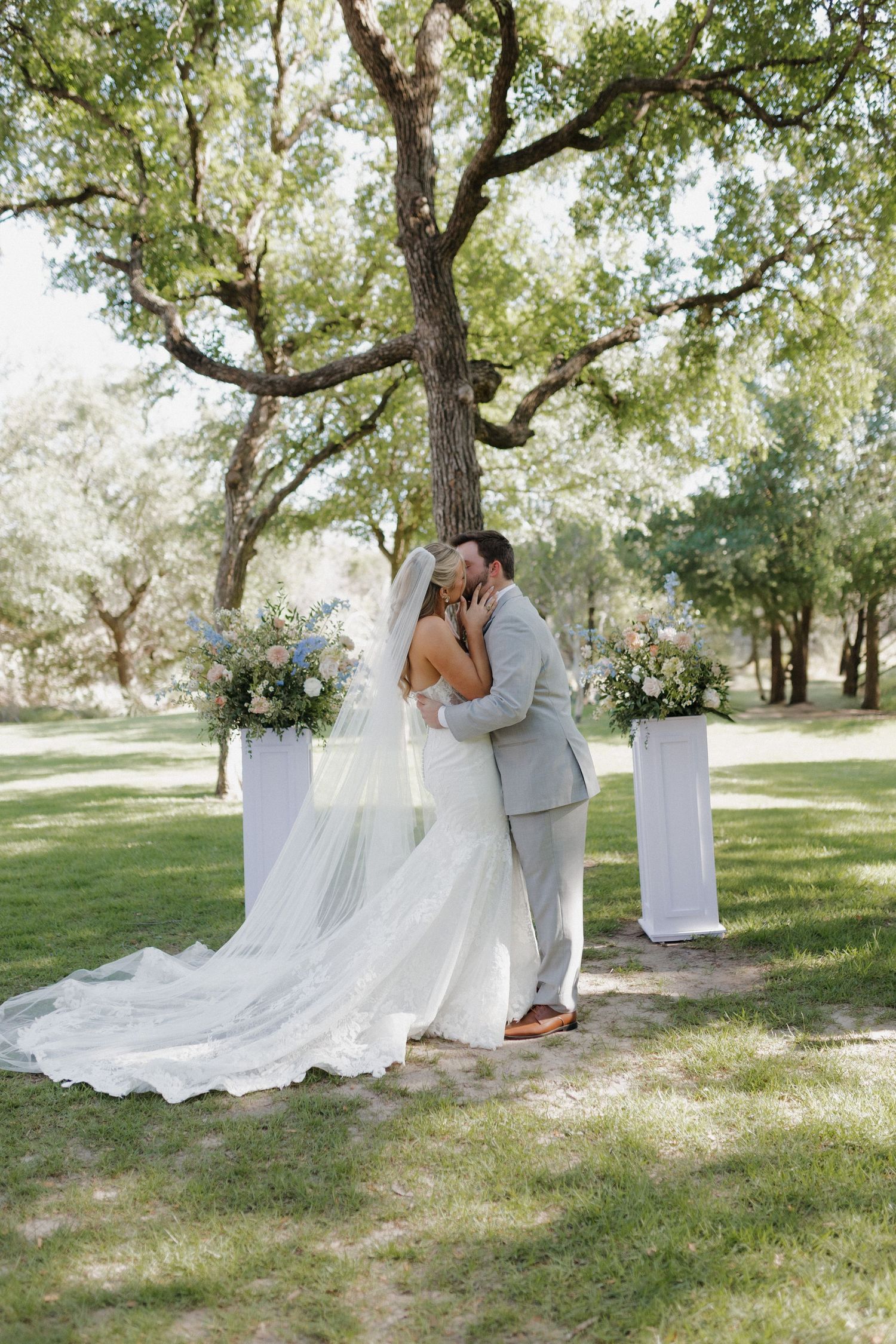 Bride and groom share their first kiss during outdoor wedding ceremony under sprawling oak trees.