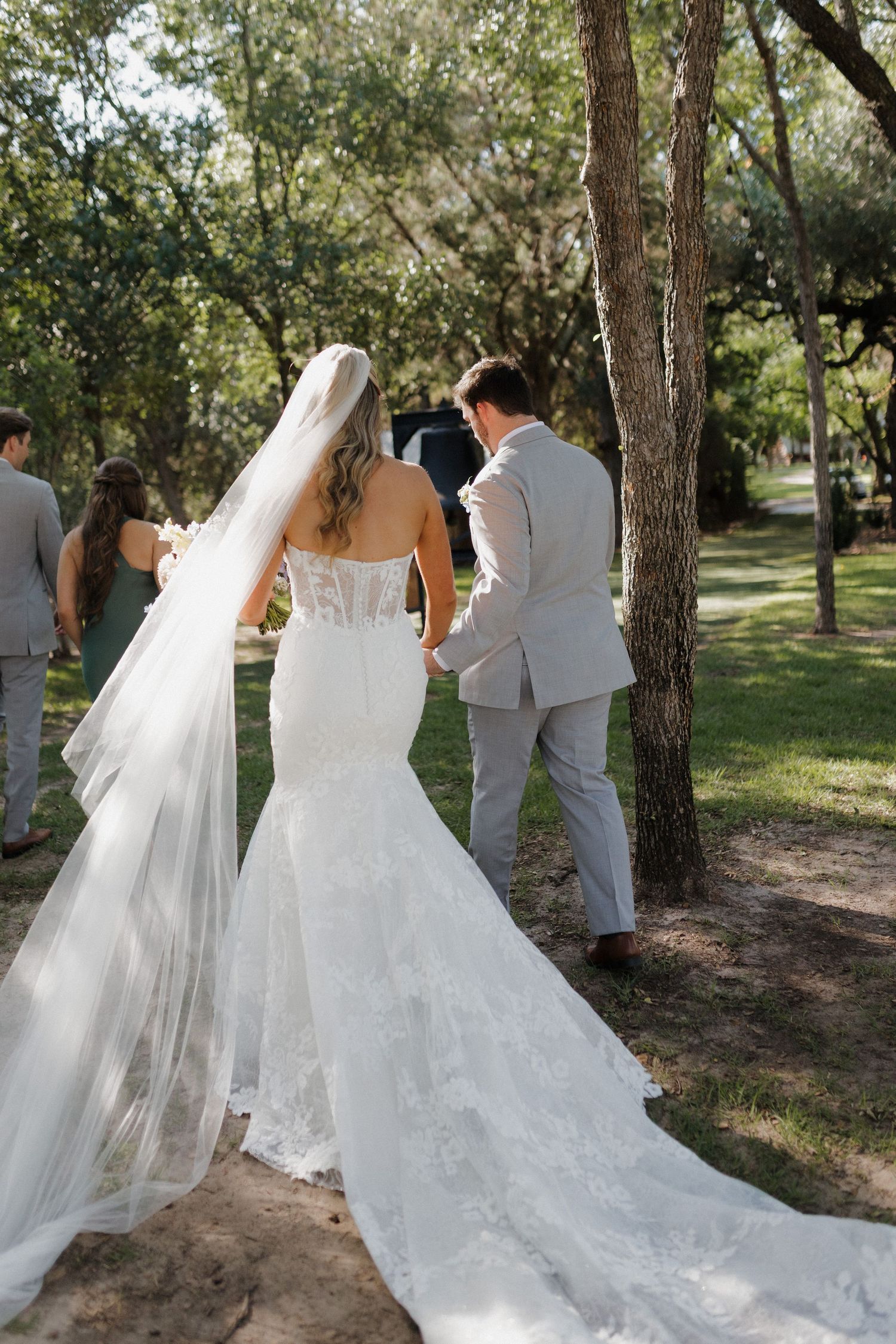 Back view of bride in lace wedding gown with flowing veil walking along tree-lined path.