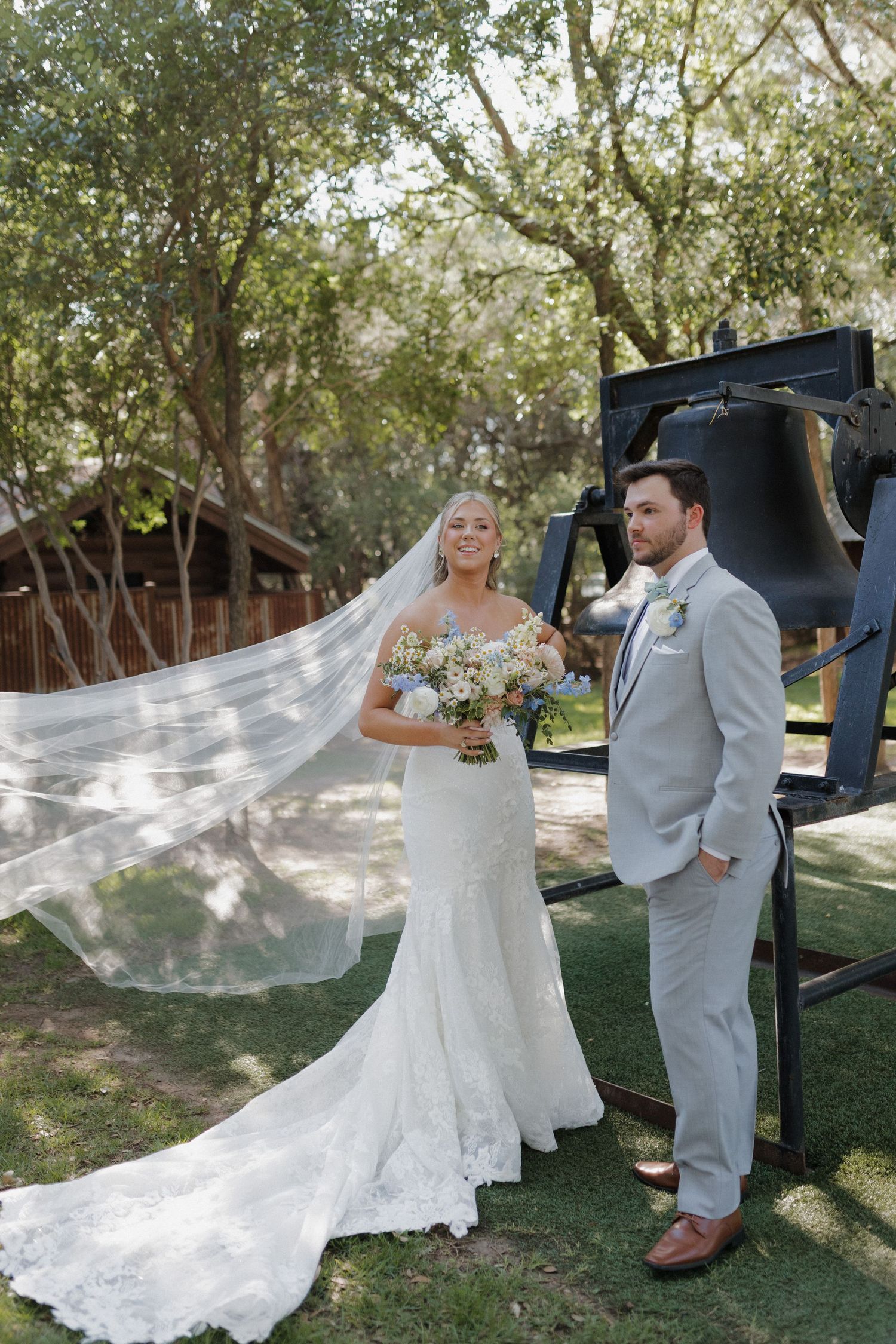 Elegant outdoor wedding portrait with flowing veil caught in breeze under shaded trees.