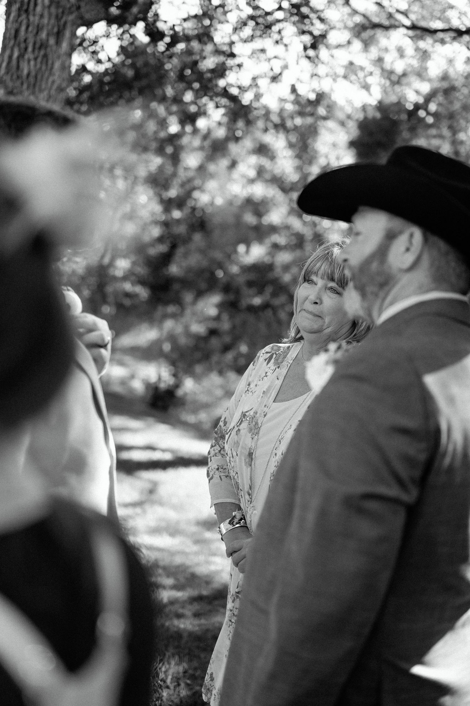 Black and white photo of a wedding ceremony gathering under trees with attendees in formal attire.