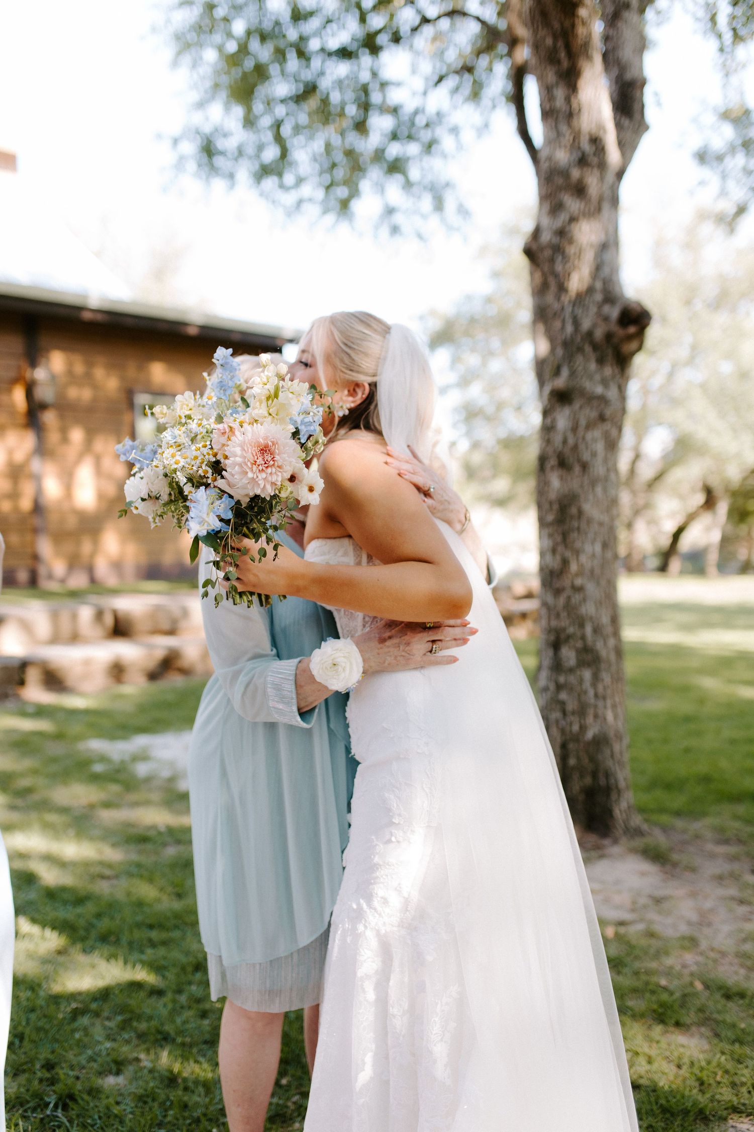 Bridesmaids in light blue dresses embracing the bride in a sunlit outdoor setting.