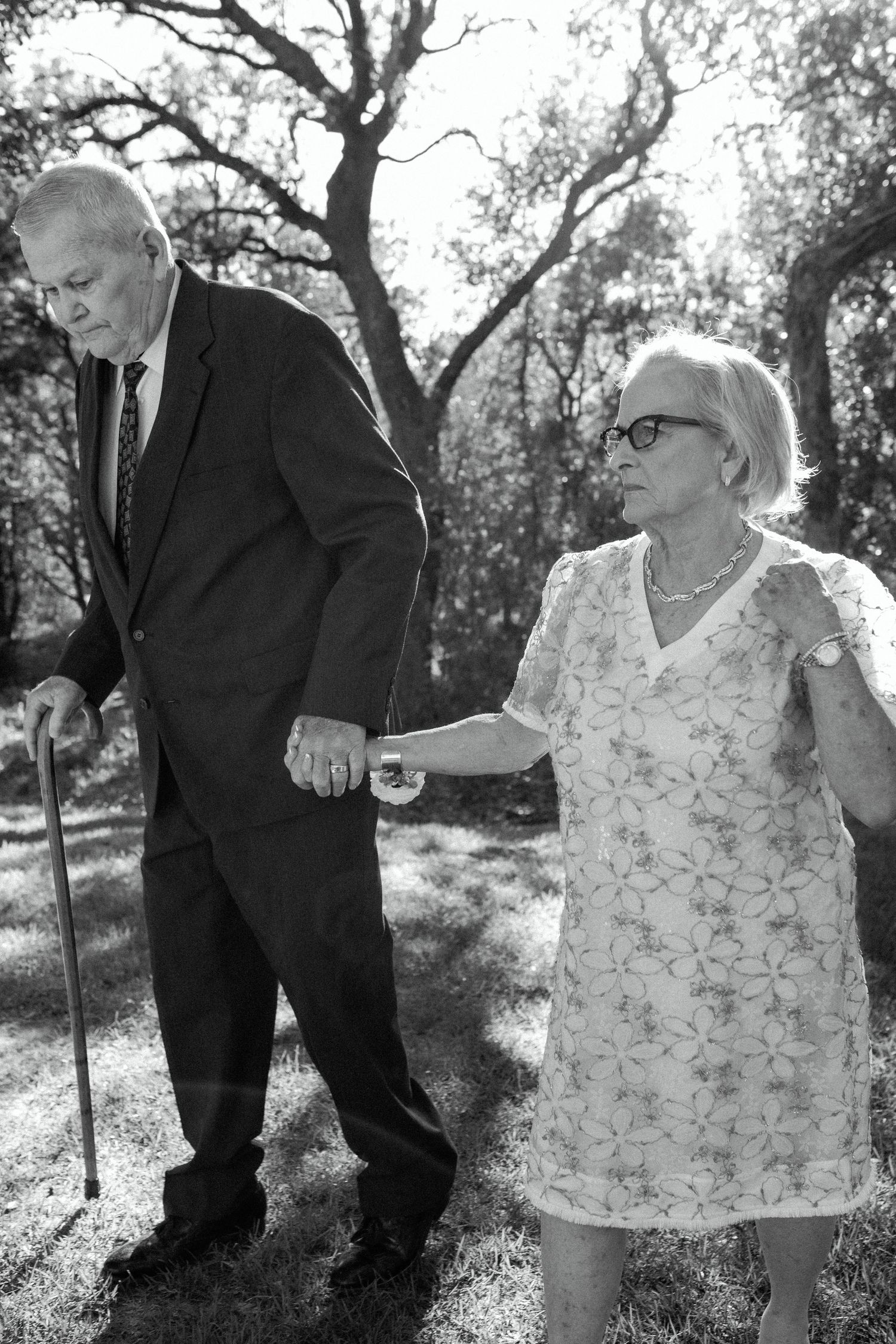 Black and white photo of elderly couple walking together at outdoor event with man using cane.