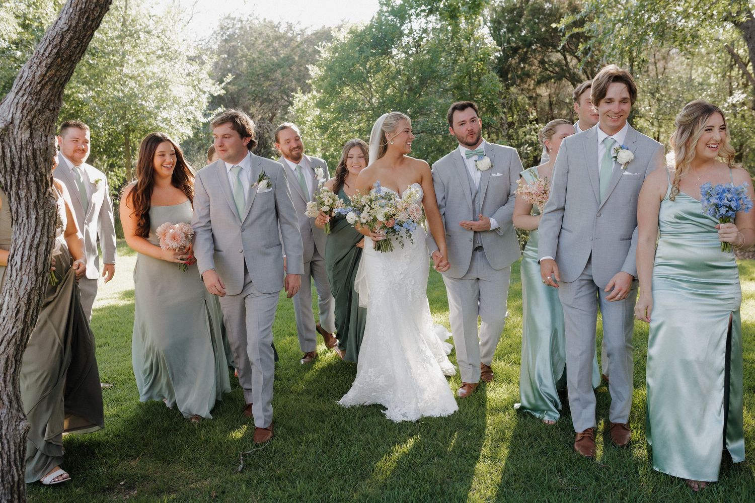 Wedding party in light gray suits and sage dresses posing together outdoors.