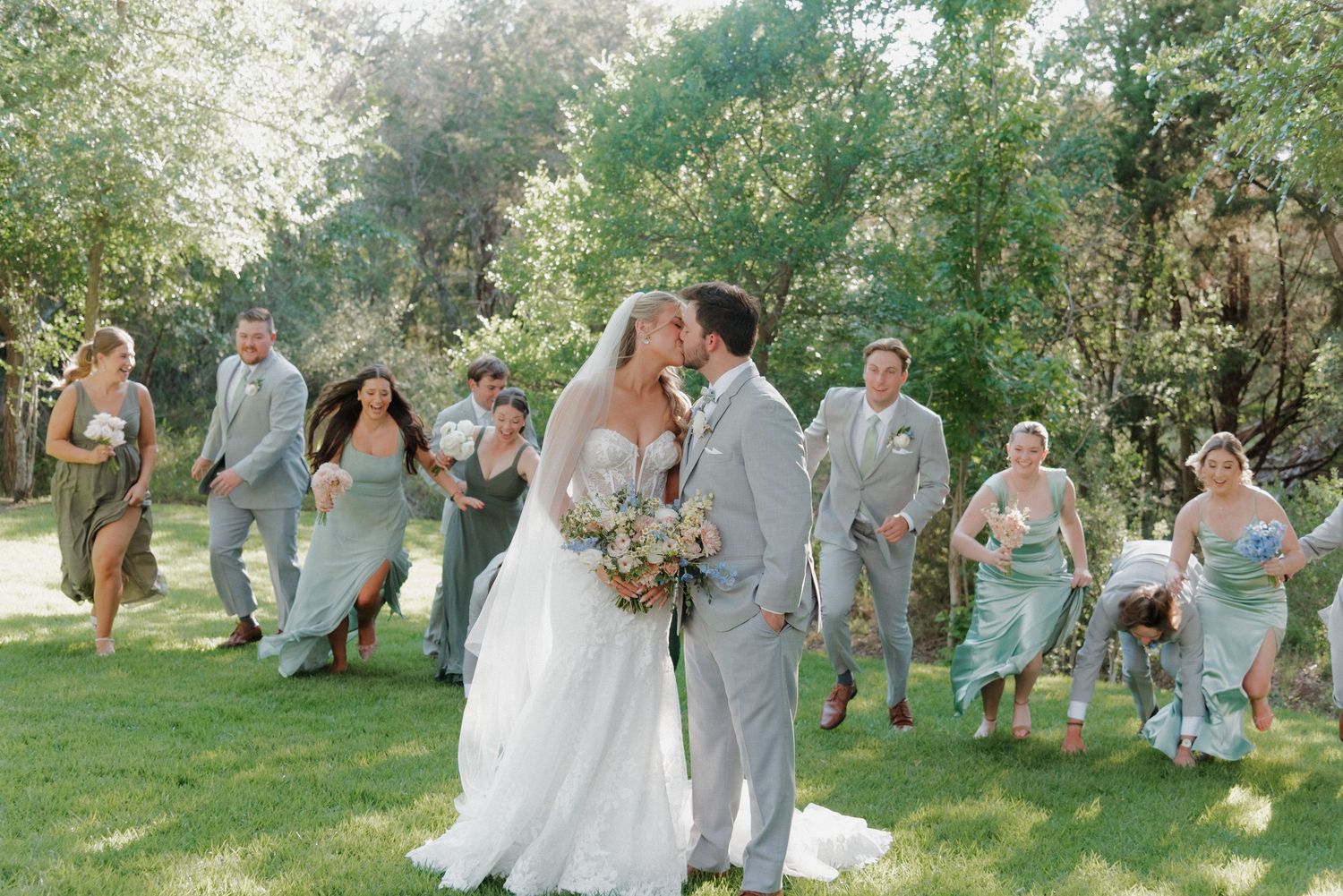 A joyful wedding party photo with people in sage green and gray attire outdoors.