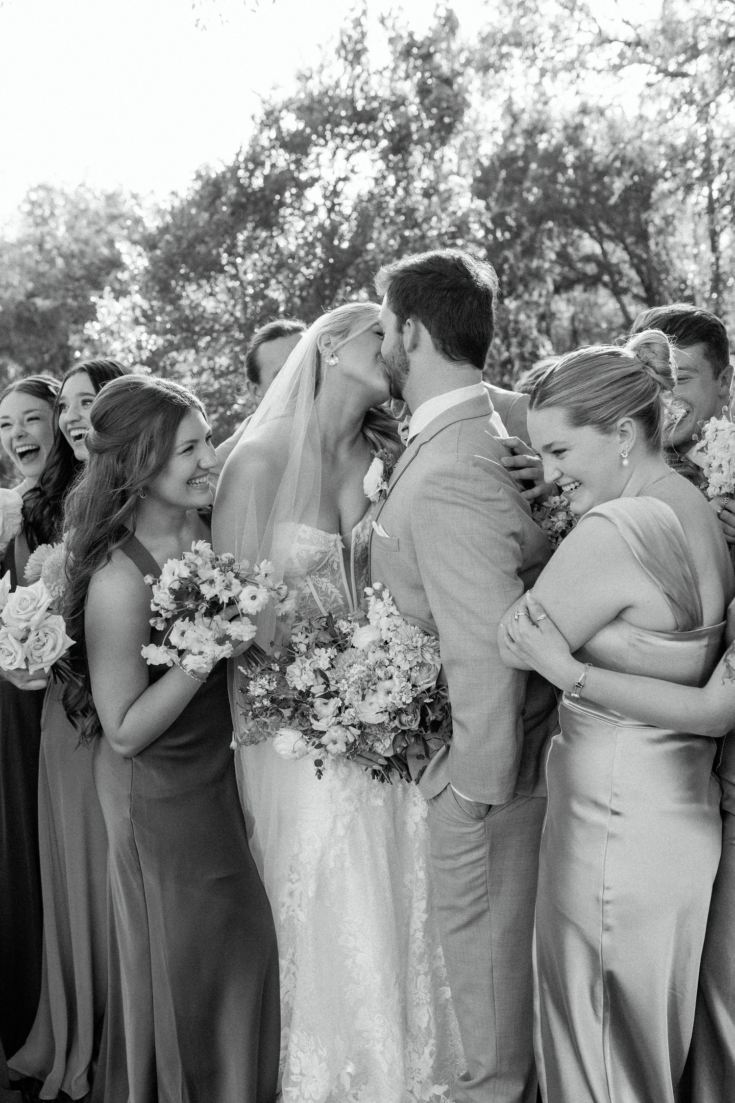 Black and white candid photo of wedding party members sharing joyful moments with bouquets.