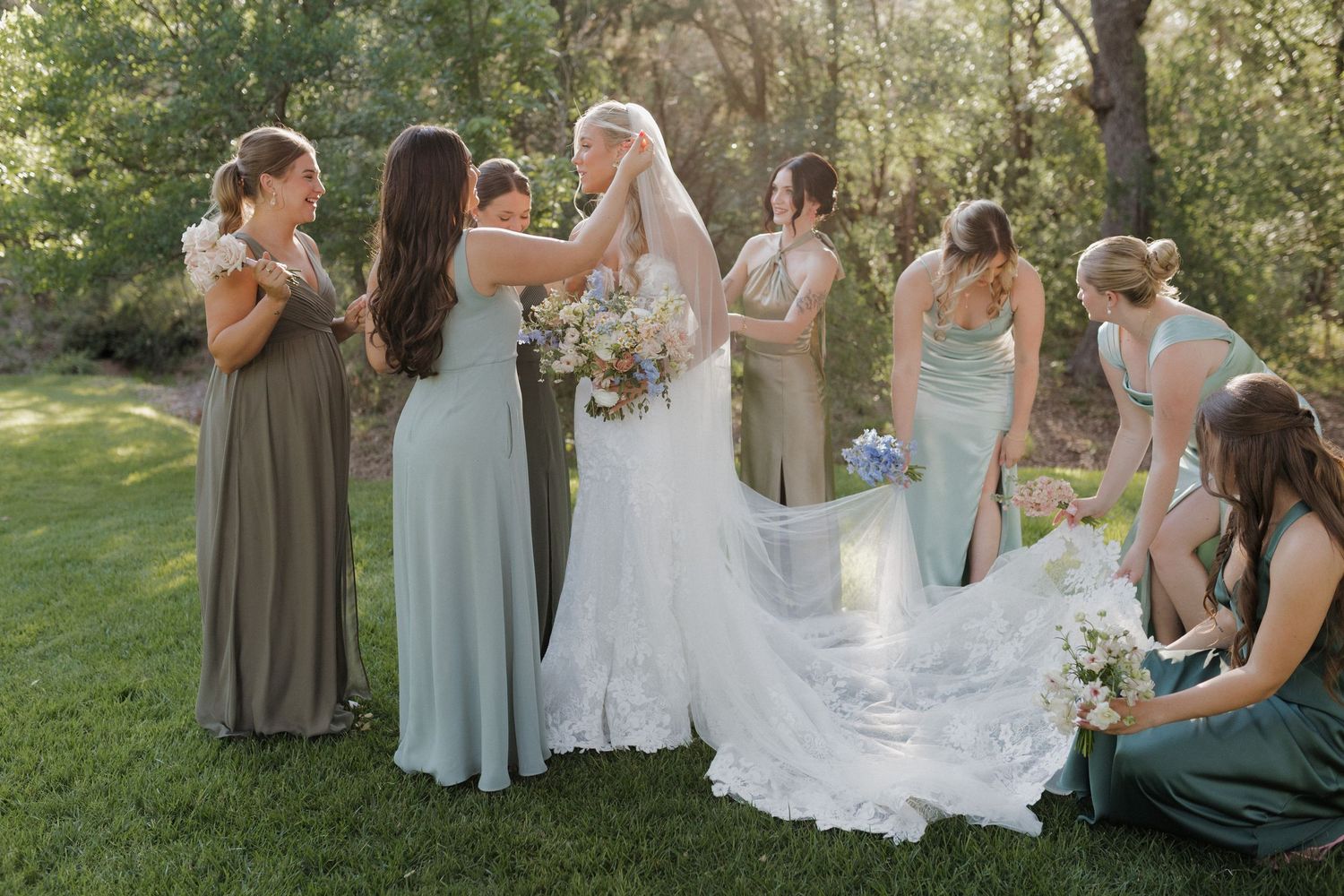Bridesmaids in soft pastel dresses helping with the bride's wedding gown outdoors.