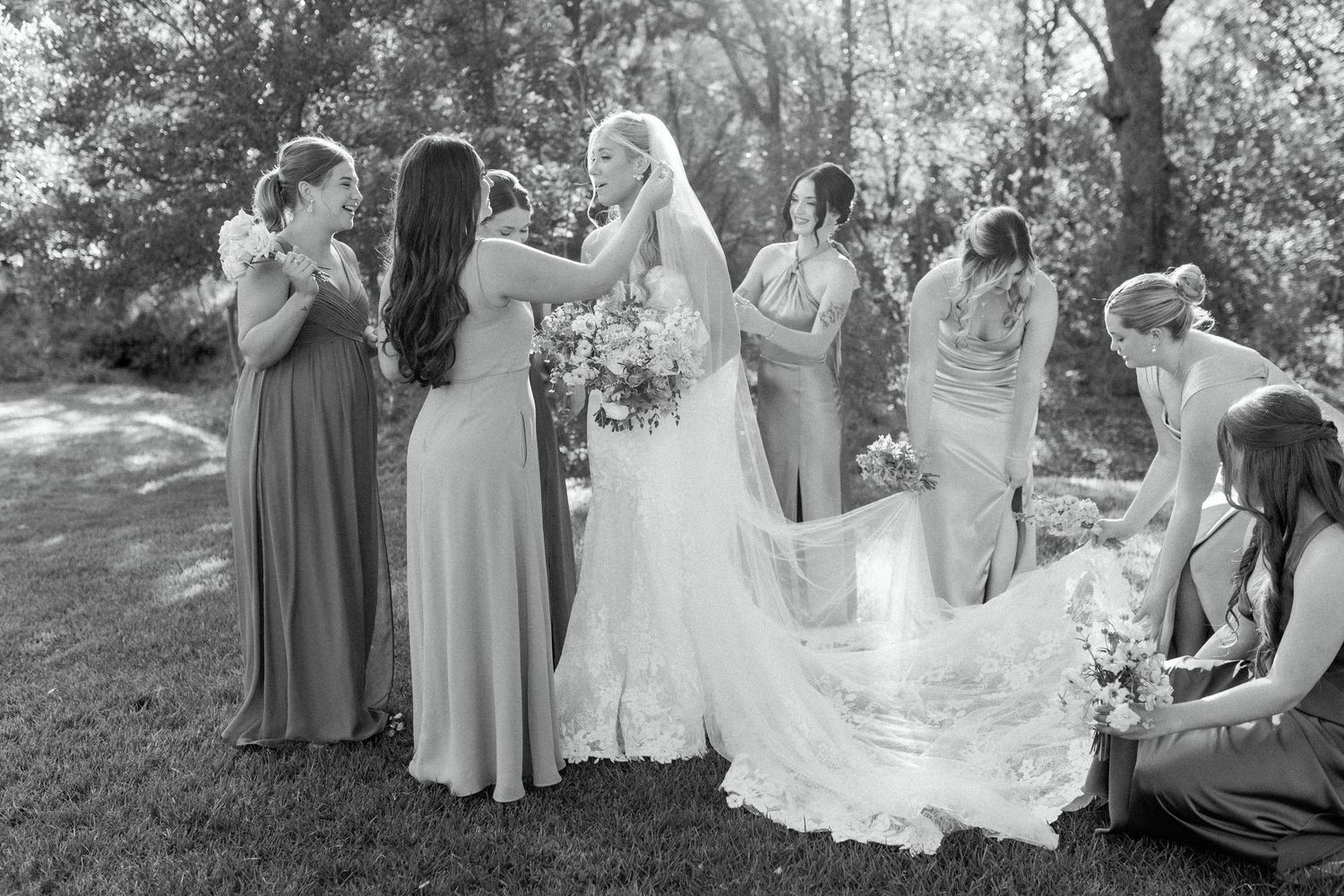 Black and white photo of bridal party helping with wedding dress and veil outdoors.