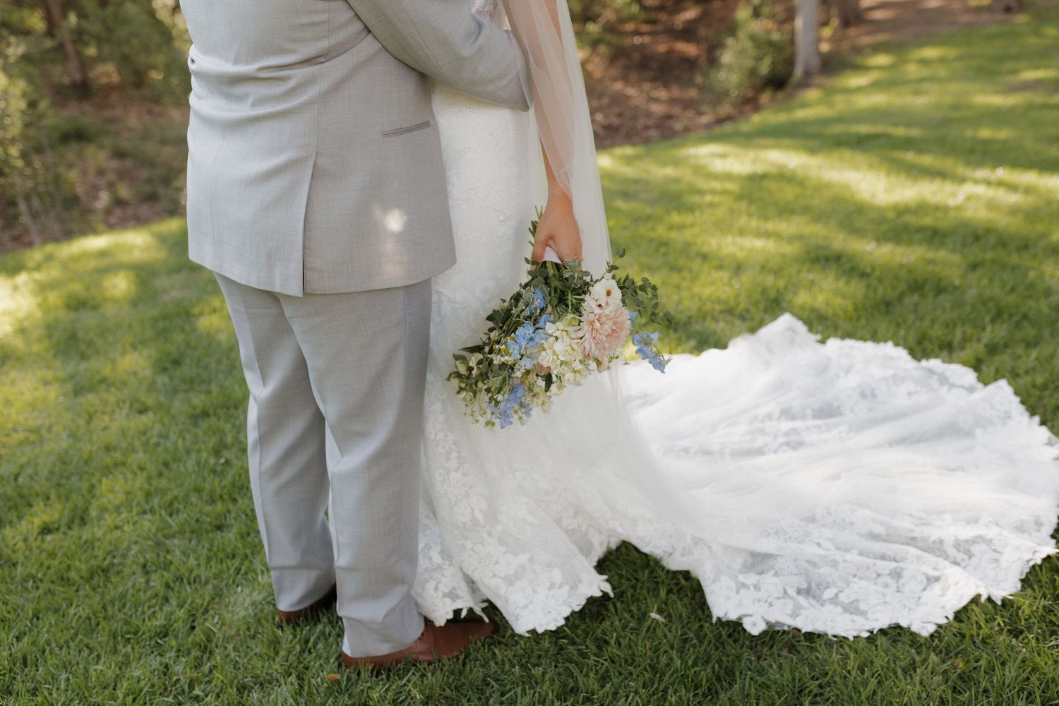 Close up view of a wedding dress train and bouquet on grass with grey suit visible.