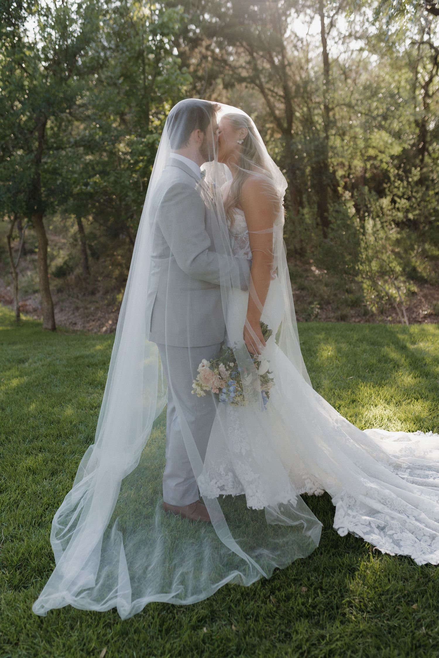 Newlywed couple in gray suit and white dress with long veil embracing on grass in sunlit garden.