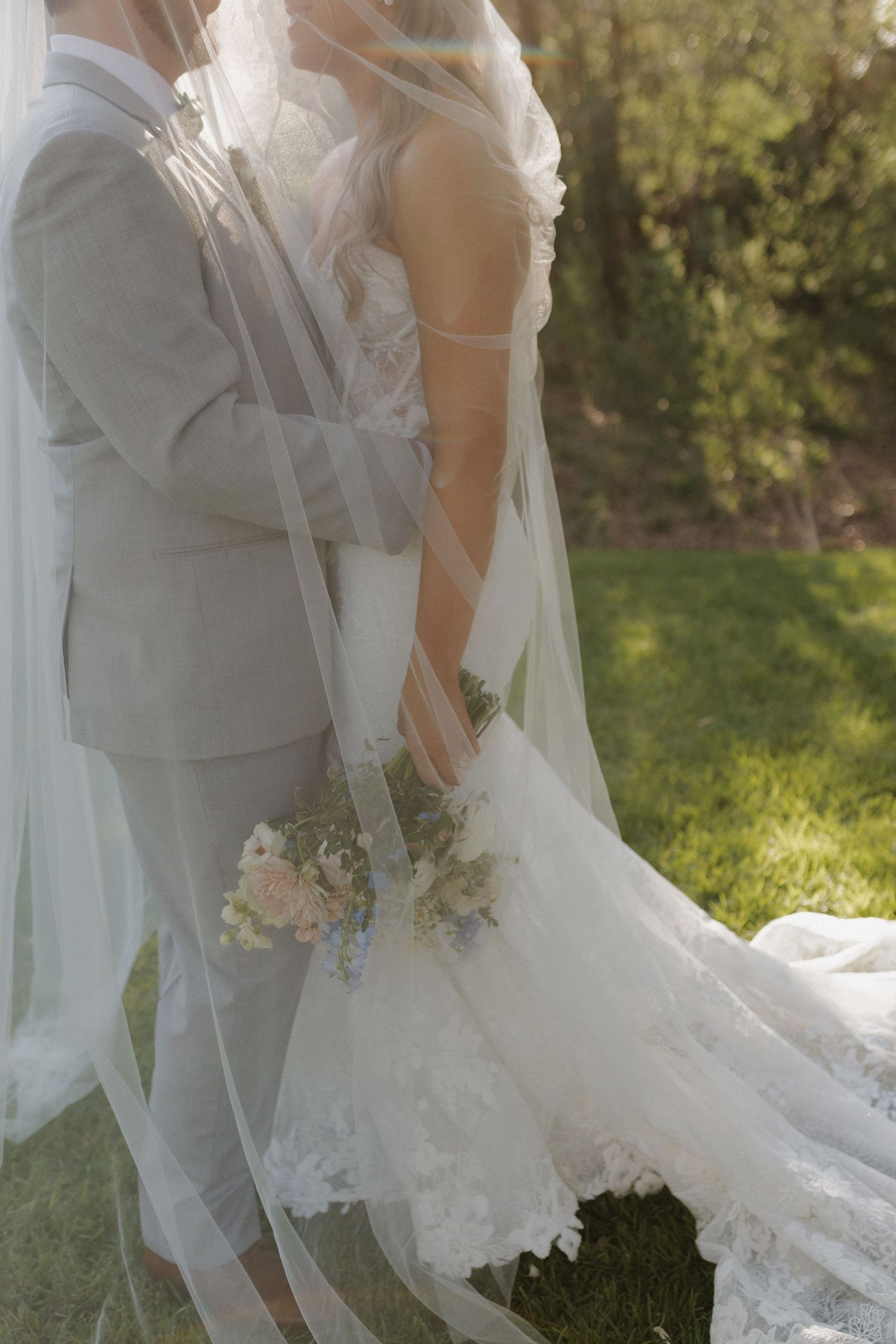 A romantic wedding photo showing the delicate details of a white lace wedding dress and gray suit in a sunlit garden setting.