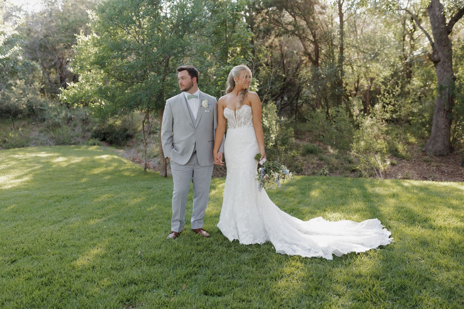 A newlywed couple stands together on a sun-lit lawn with the bride wearing a flowing white gown.