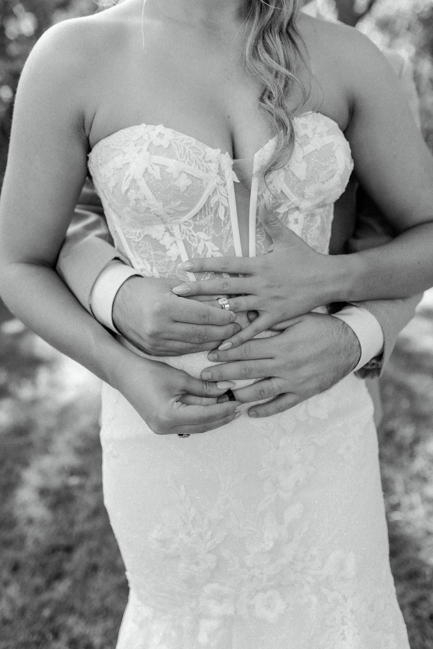 Close-up black and white shot of a bride's torso in a detailed lace wedding dress with hands embracing.