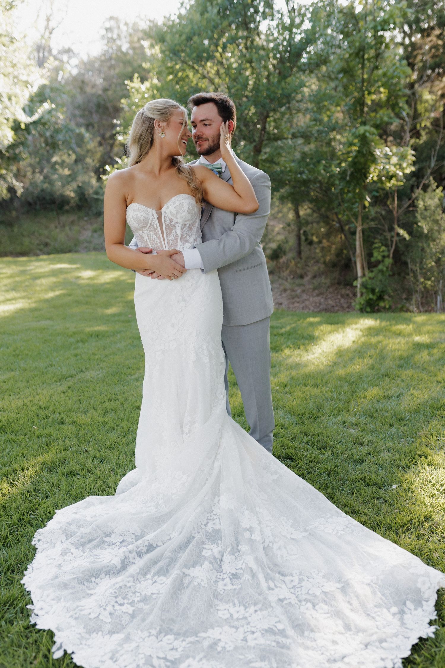 A romantic wedding portrait showcasing a bride's detailed lace train as couple embraces on garden lawn.