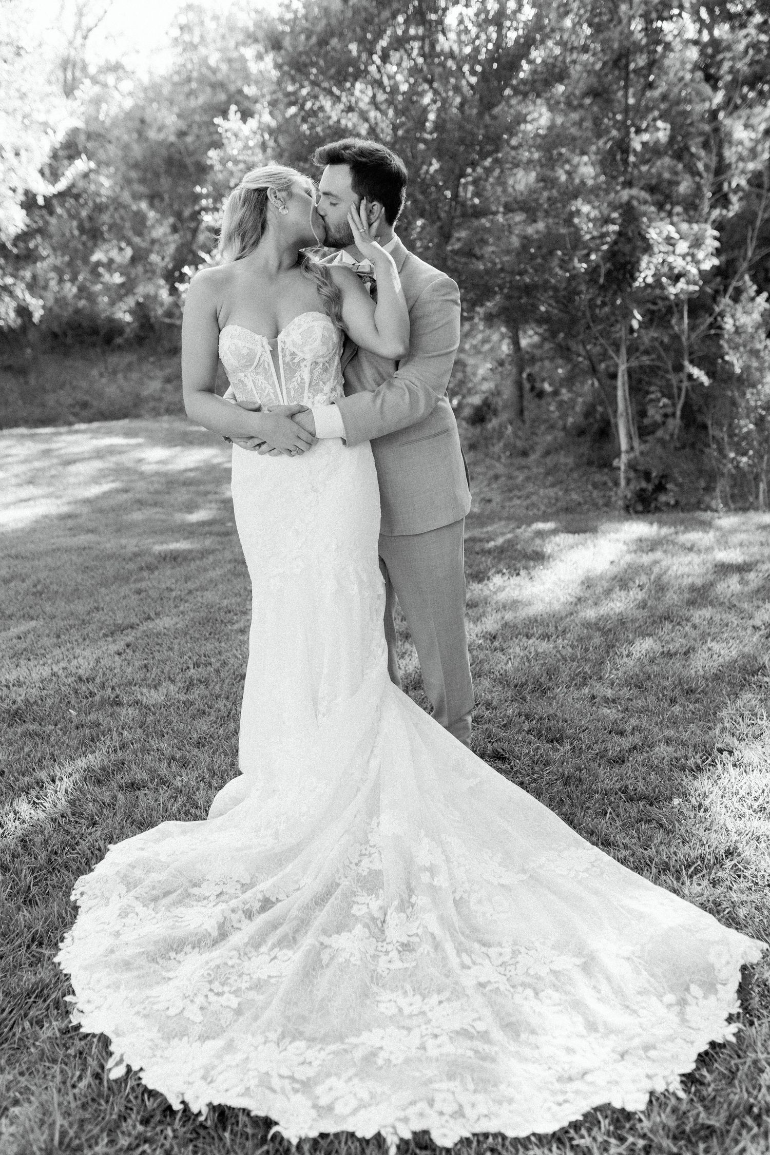 Black and white portrait of newlyweds embracing, showcasing bride's detailed lace train on outdoor lawn.