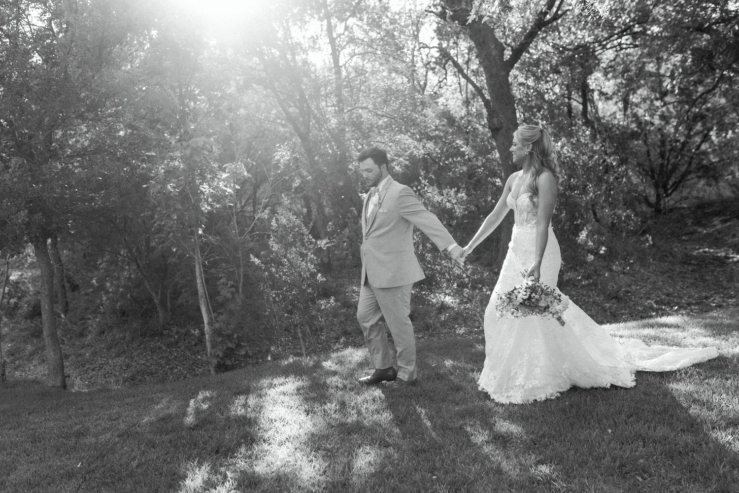 A black and white photo of newlyweds walking hand in hand through a sunlit garden setting.
