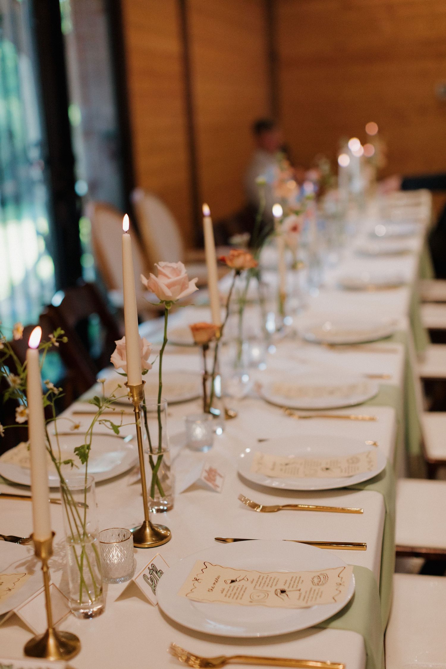 Elegant wedding reception table setting with white plates, brass candlesticks, and delicate floral centerpieces in glass vases.