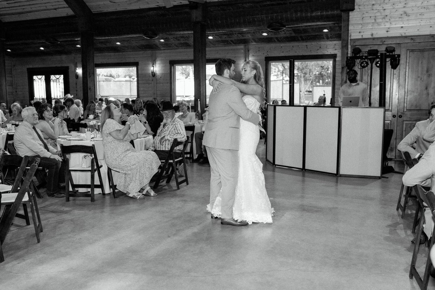 Black and white photo of a couple sharing a dance in rustic reception venue with guests seated nearby.