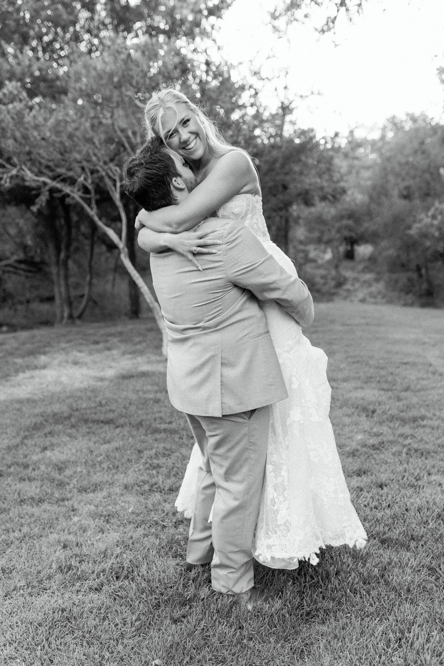 A joyful black and white wedding photo showing a couple embracing on a grassy lawn surrounded by trees.