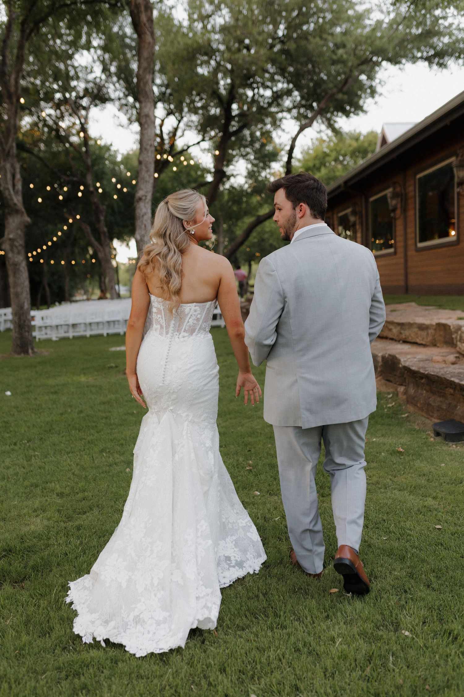 Bride in fitted lace wedding gown and groom in gray suit walking together on lawn with string lights and cabin.