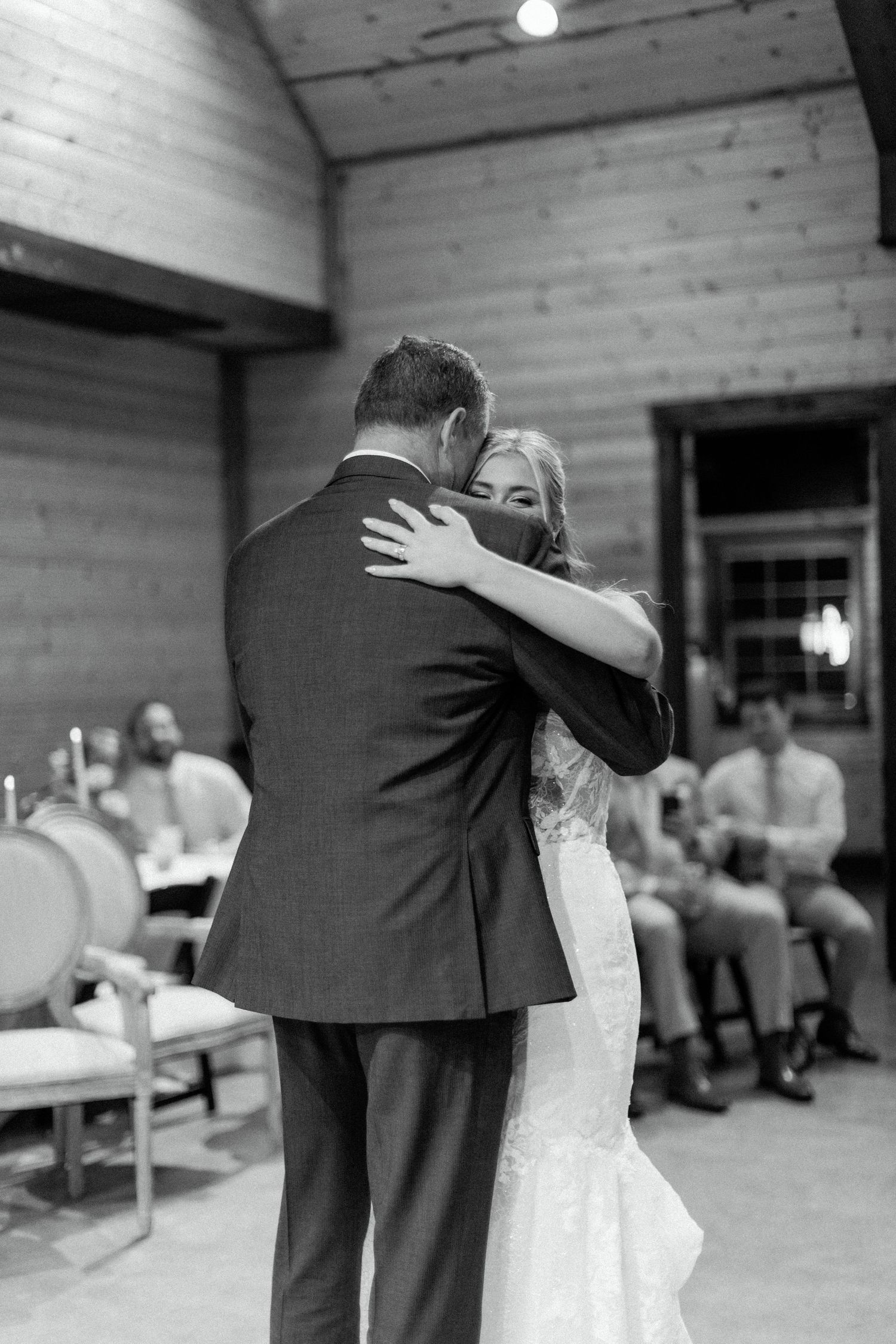 A couple shares their first dance at a rustic barn wedding reception while guests look on in black and white.