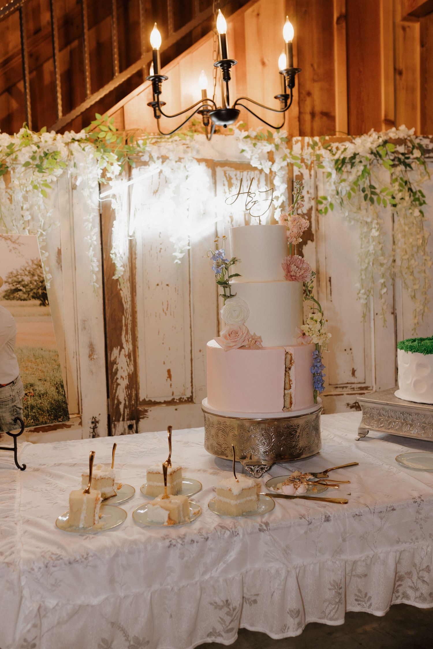 Elegant three-tiered white wedding cake displayed on rustic table with candlelit chandelier and cascading floral decorations.