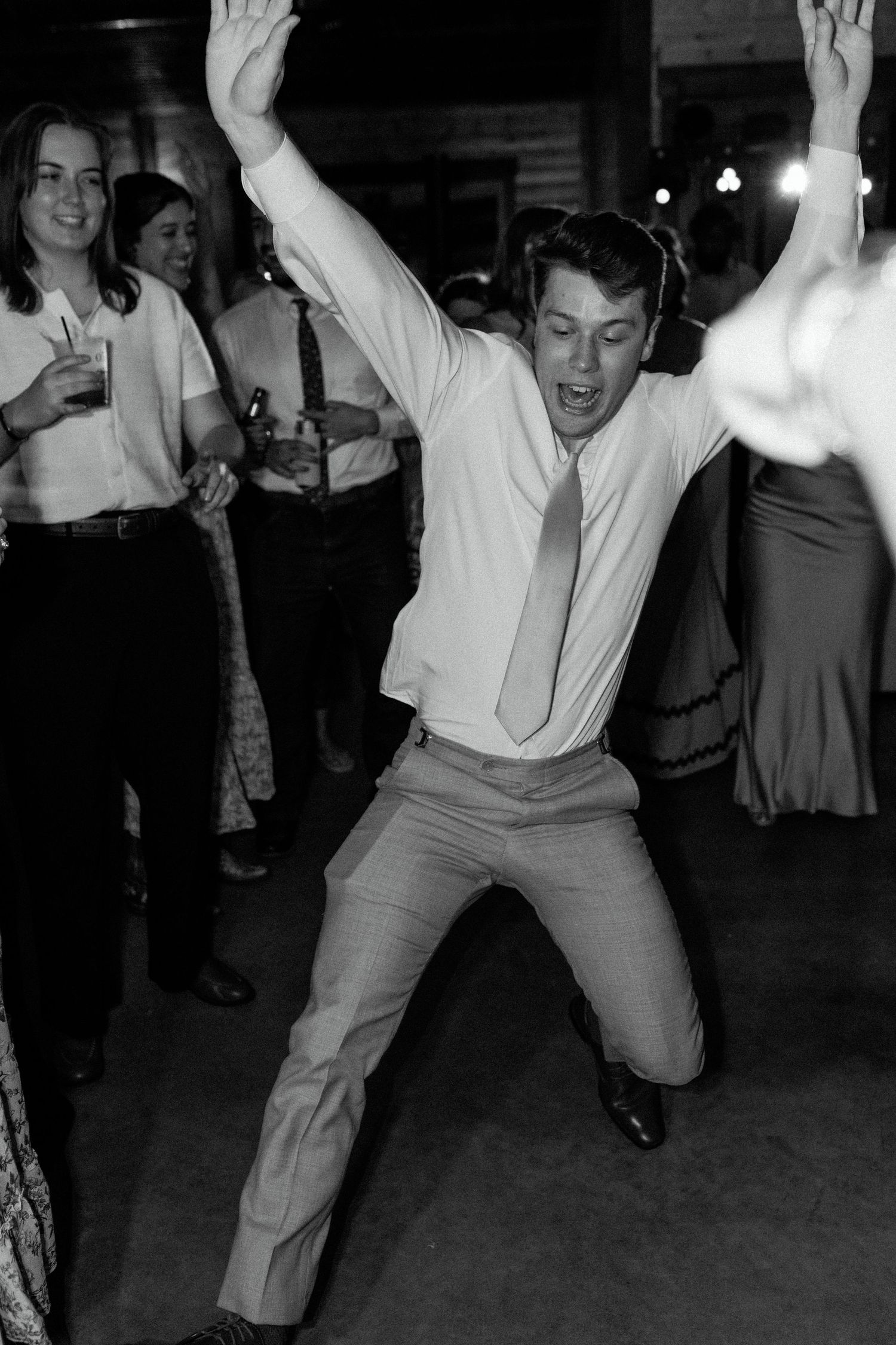 A person dances energetically on a dance floor in a white button-down shirt and tie at what appears to be a vintage party.