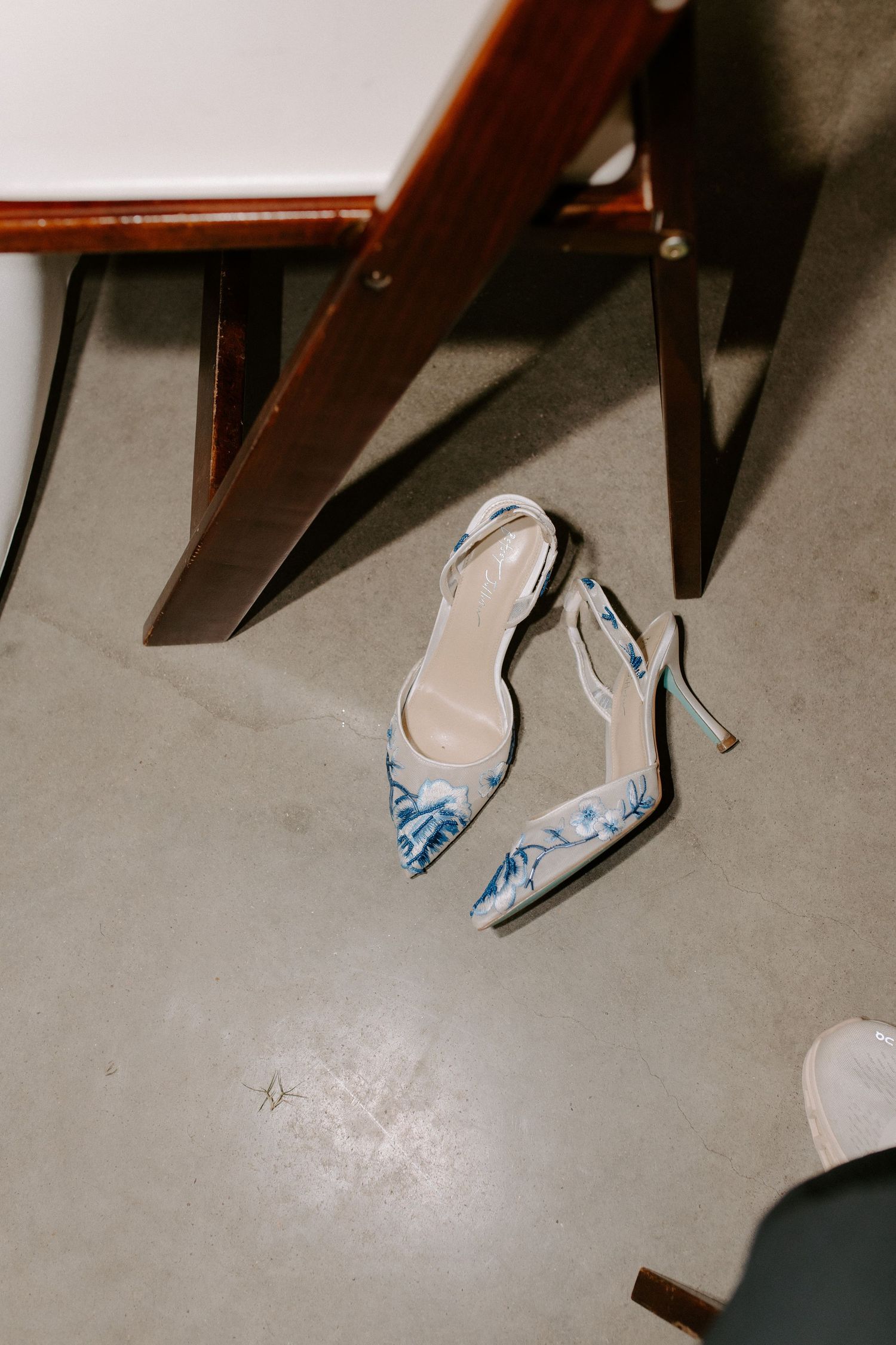 Elegant blue and white floral slingback heels with pointed toes rest on a grey floor beneath wooden furniture.