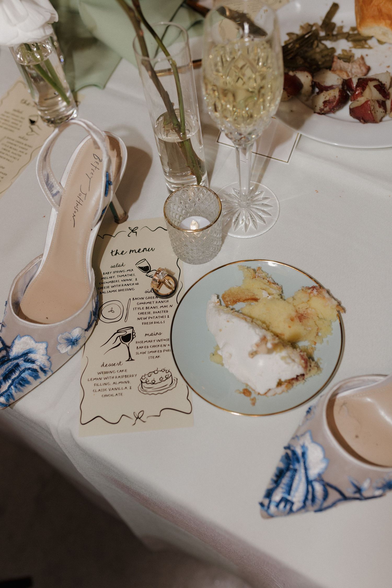 A table setting with a champagne glass, white heel shoe, menu card, and partially eaten dessert on a blue and white tablecloth.