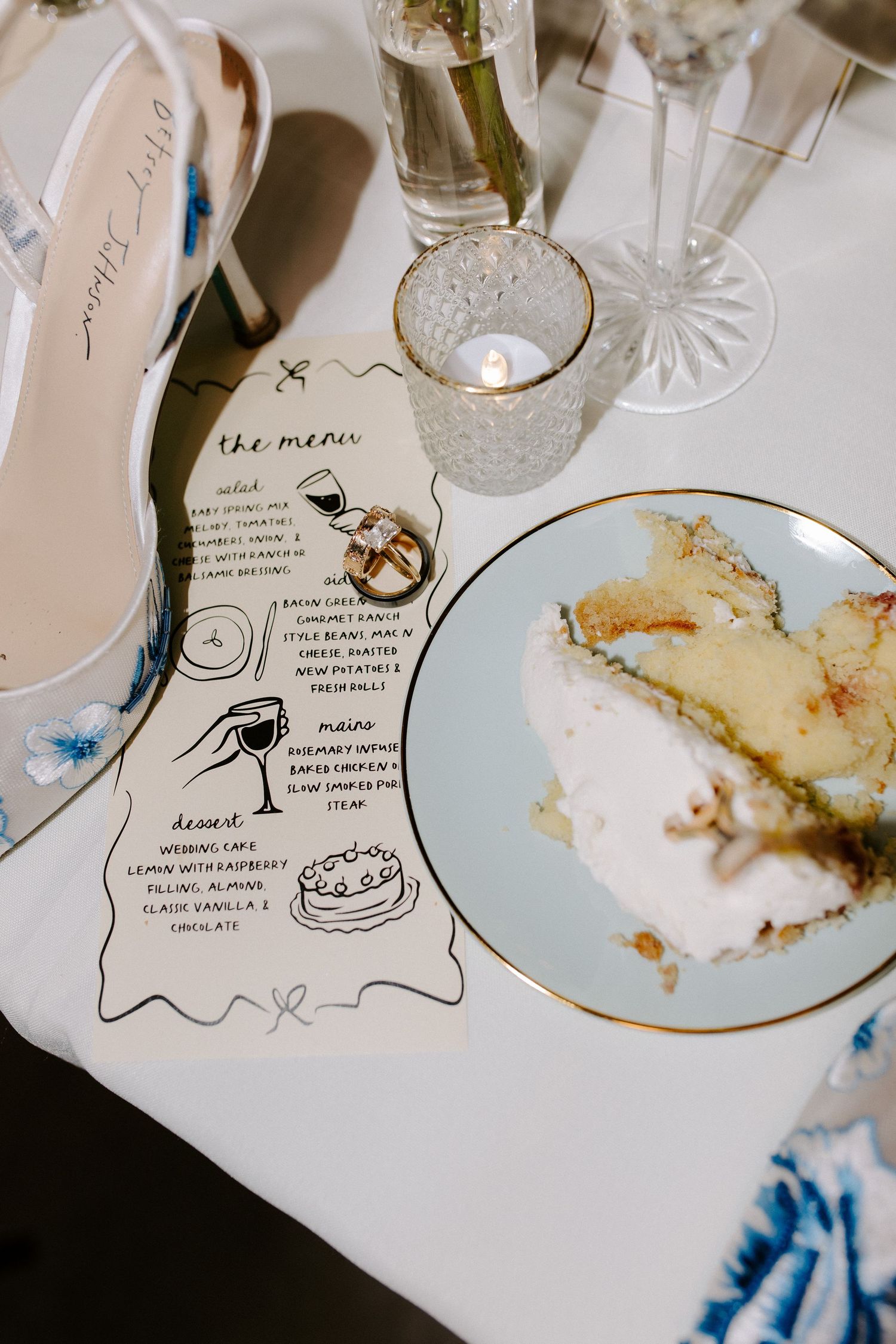 Hand-drawn menu card placed beside an elegant shoe, crystal glass and remnants of dessert on a white plate with gold rim.
