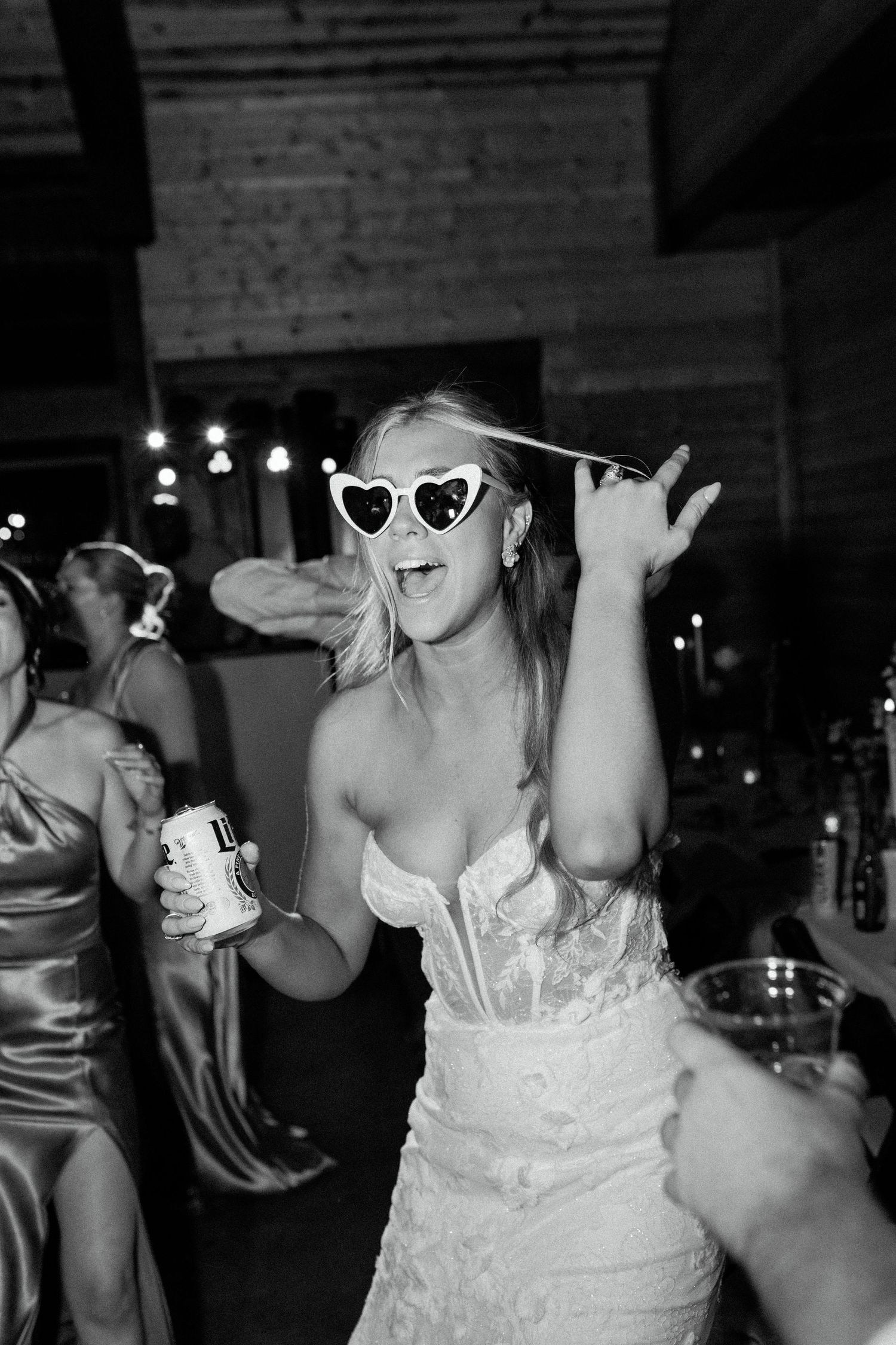 A person in a white lace wedding dress dances joyfully wearing heart-shaped sunglasses at a reception.
