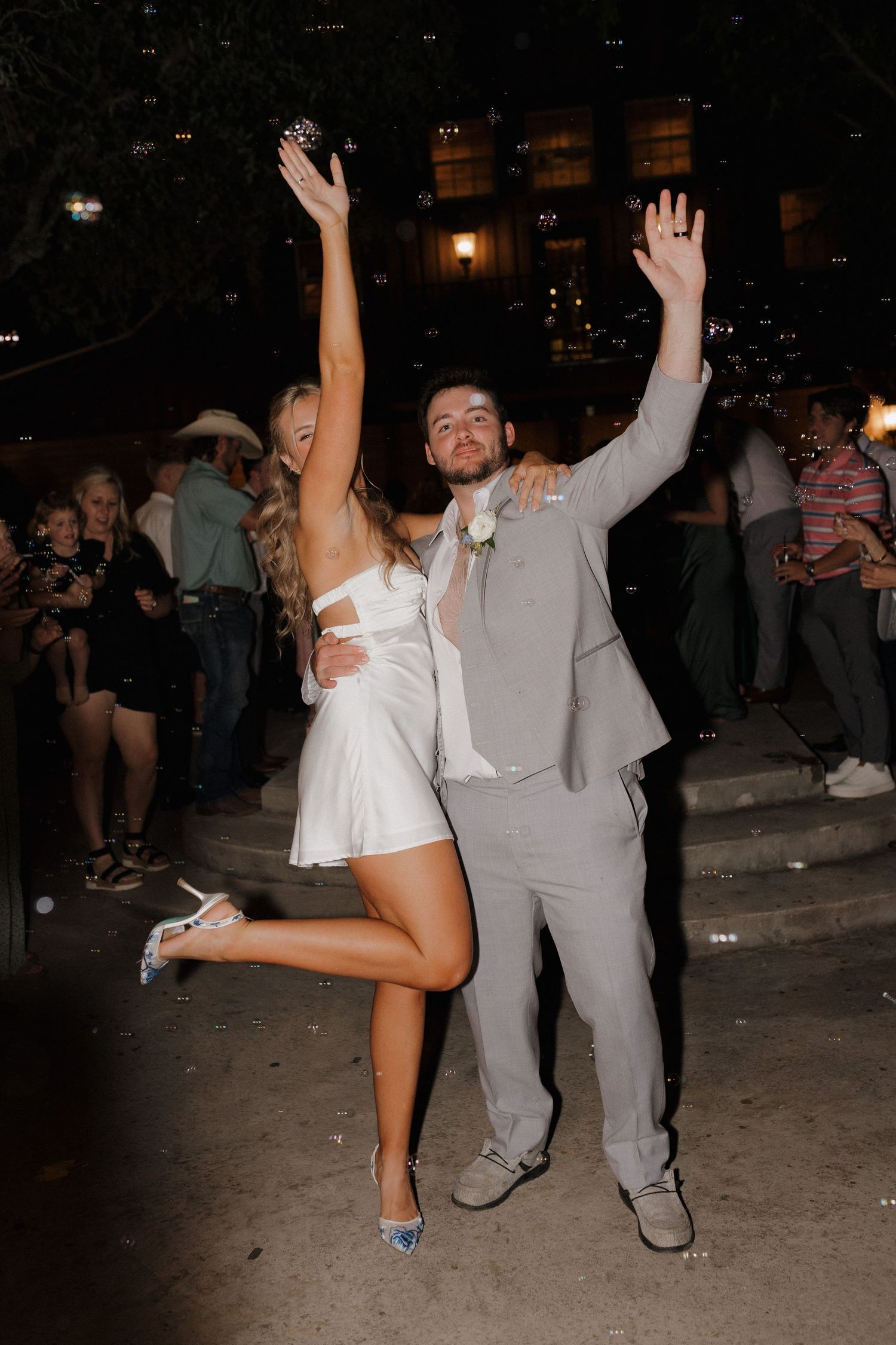 A joyful couple dancing at a wedding reception, with the bride in a short white dress and groom in gray suit.