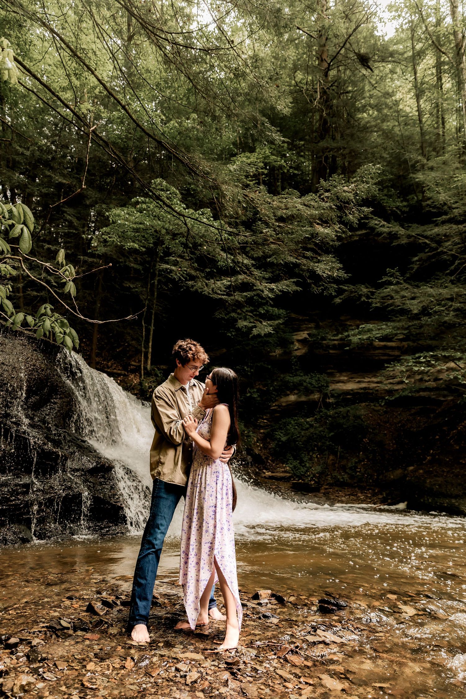 Couple embraces near scenic waterfall in lush forest creek during romantic outdoor photoshoot.