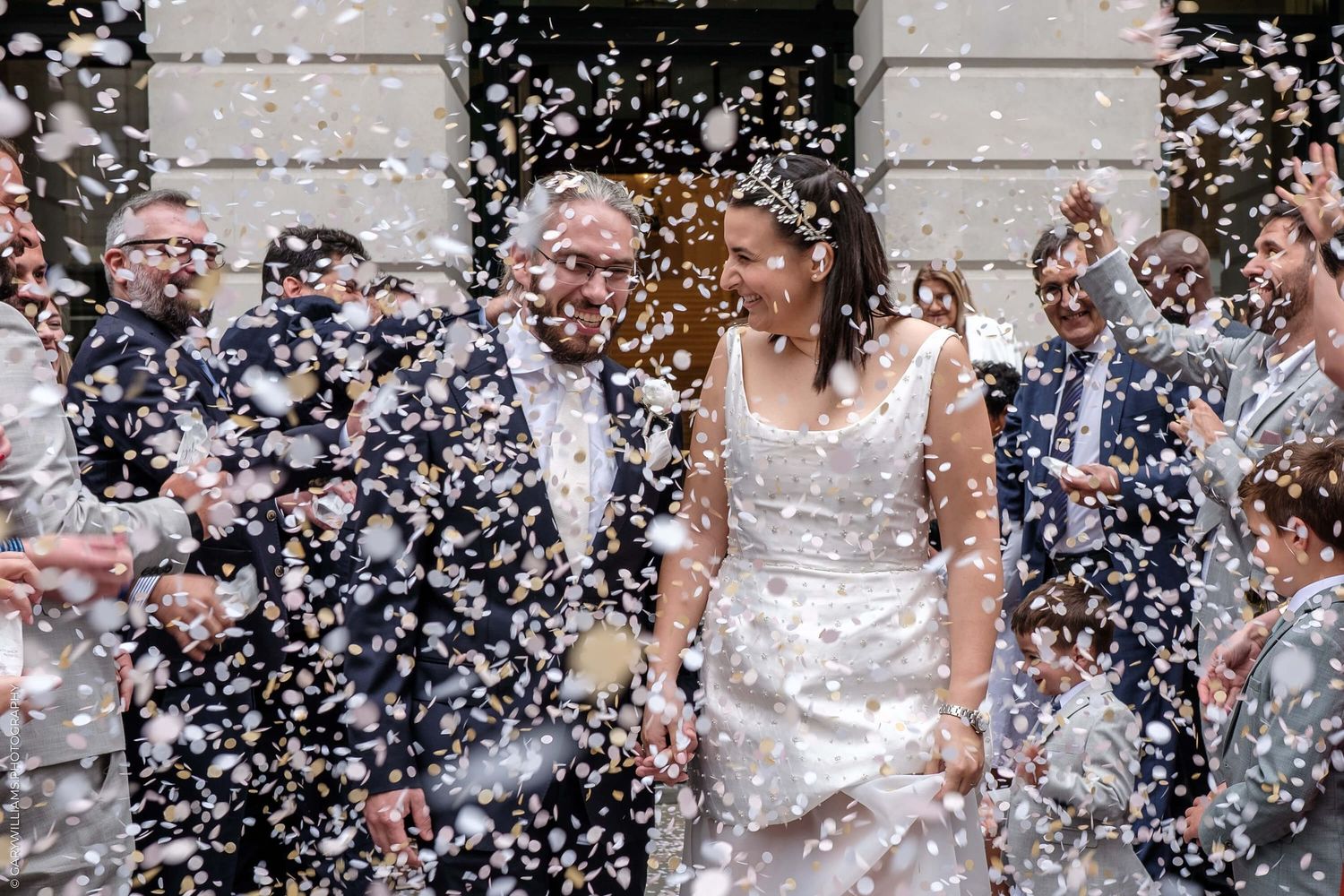 Wedding celebration with confetti raining down on newlyweds and guests outside a grand stone building.