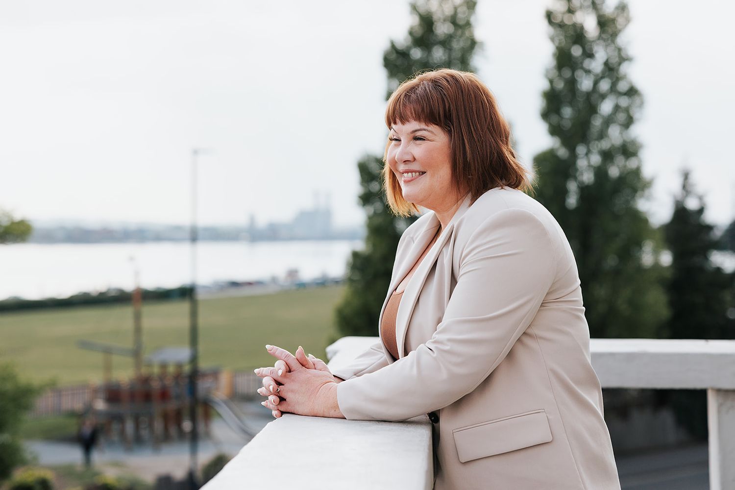 A smiling individual wearing a beige blazer leans against an outdoor railing overlooking a scenic waterfront view in Southampton