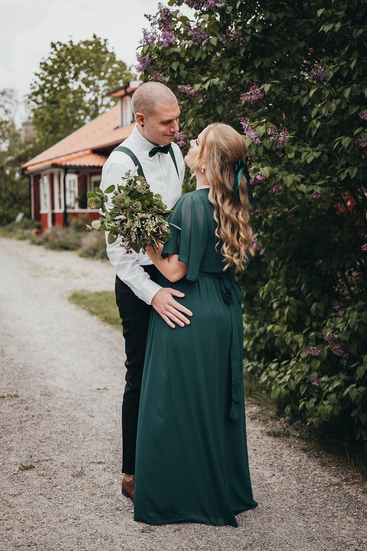 Bröllopspar står intill en blommande syrenbuske i lantlig miljö med ett rött trähus i bakgrunden vid Borghult Lapphem.