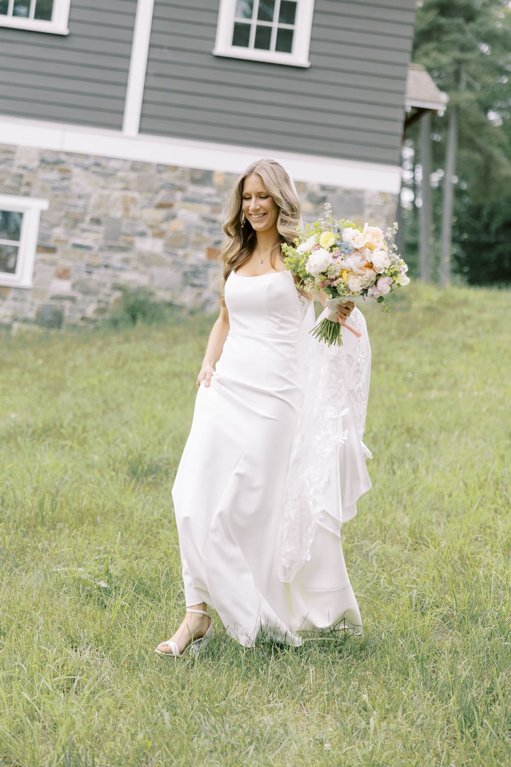 A bride in a flowing white wedding dress holds a pastel bouquet while walking through a grassy field near a stone house.