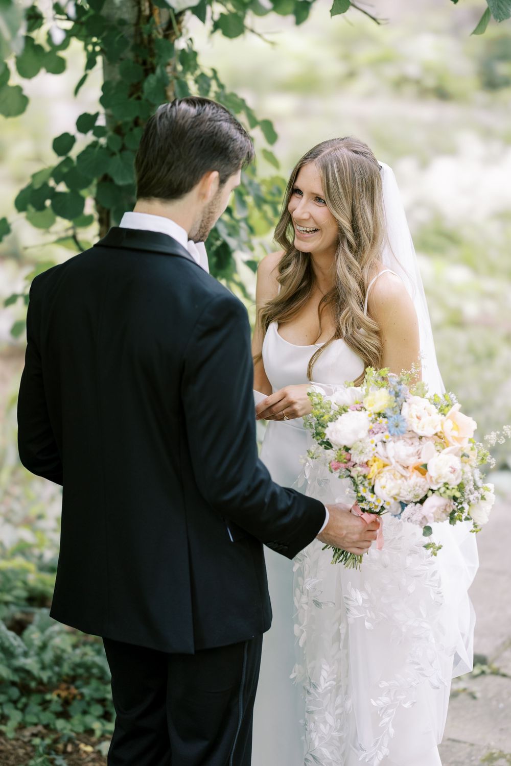 A couple in wedding attire share a joyful moment outdoors, with the bride holding a romantic pastel floral bouquet.