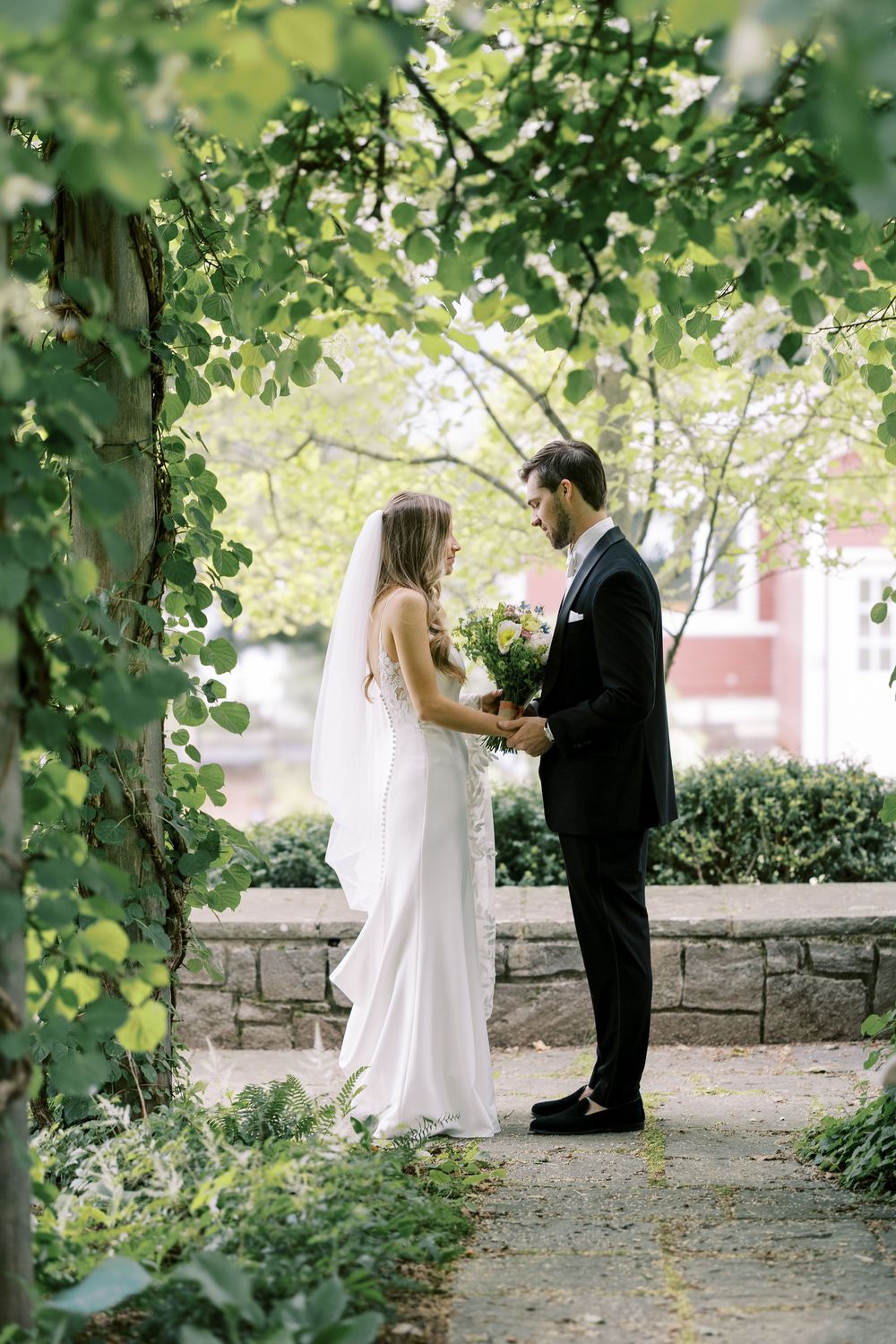 A couple stands together under leafy green trees on a stone-lined garden path during their wedding ceremony.