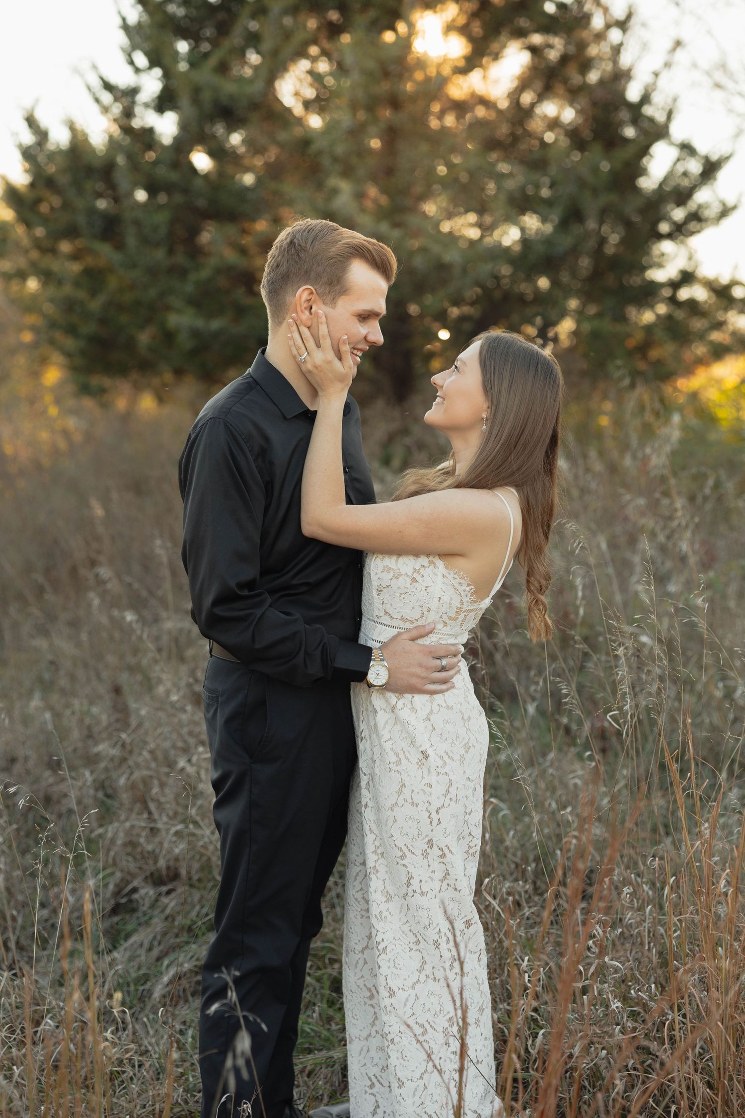 A couple embraces in a romantic outdoor setting at sunset with tall grass and evergreen trees in the background.