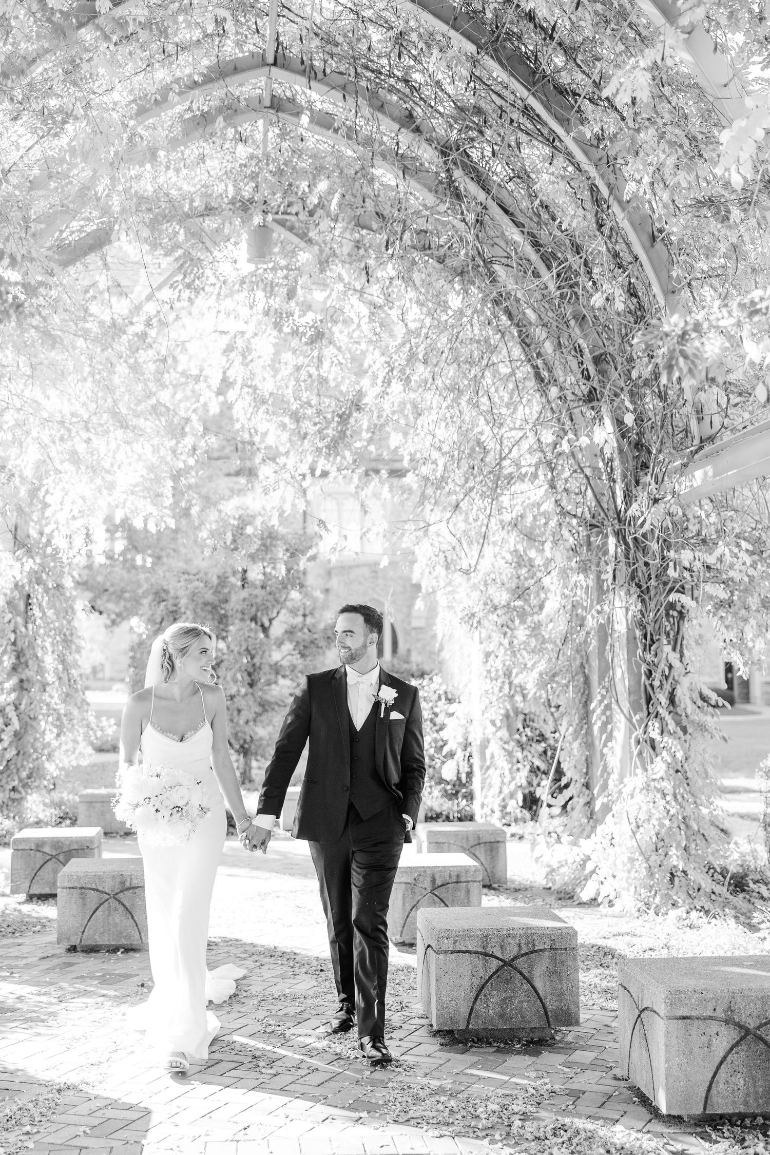 A romantic couple walking through a vine-covered garden archway in wedding attire.